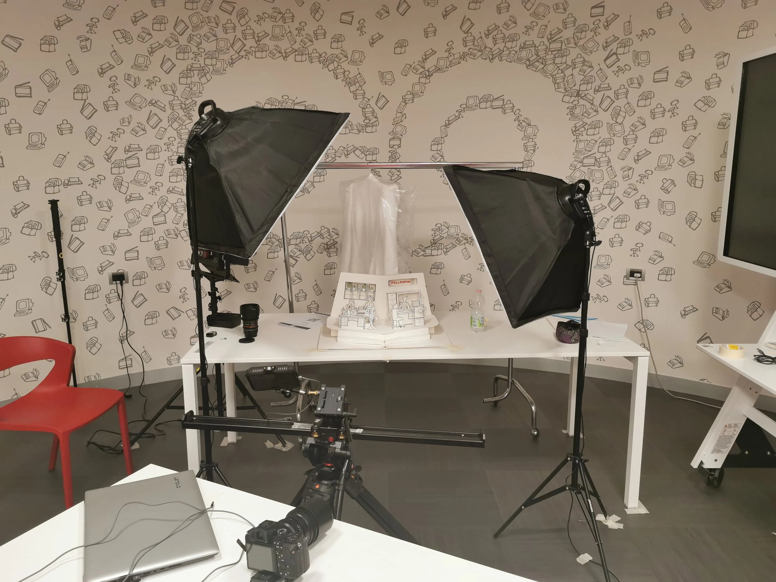 A photography or video studio setup with a white table in front of a patterned wall, two large softbox lights on stands aimed at the table, a camera on a slider, and a camera on a tripod. On the table are some sketches, a water bottle, a lens, and a