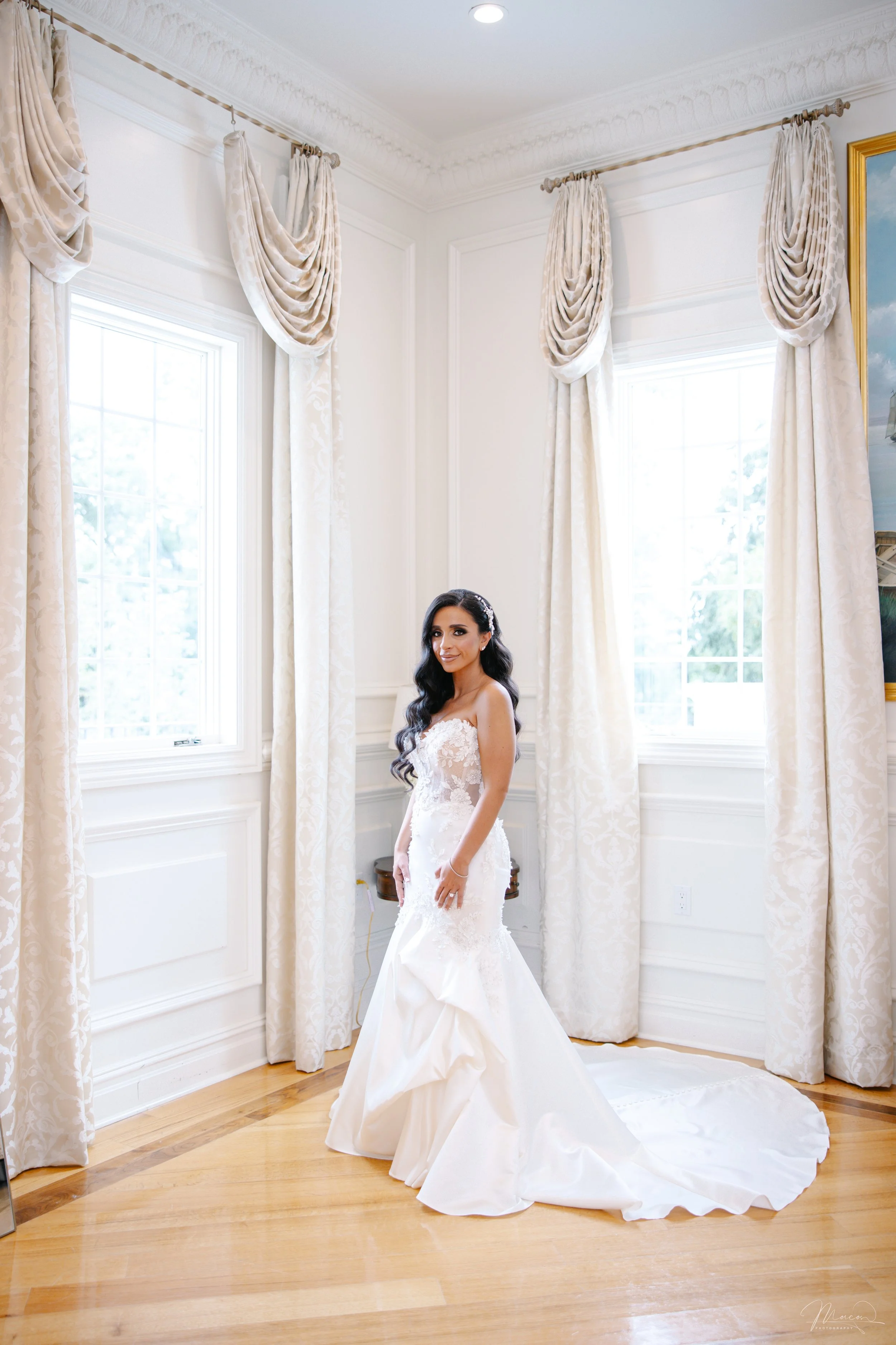 A bride in a white wedding dress standing in a well-lit room with white walls, large windows, and ornate white curtains.