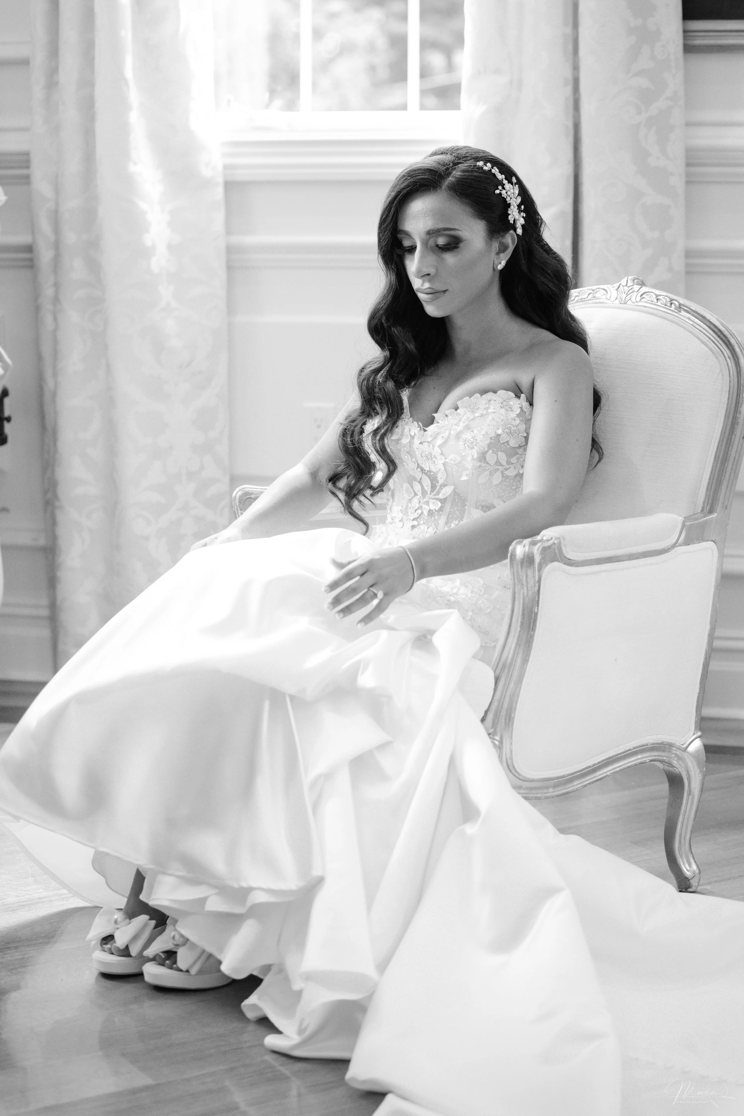 Black and white photo of a woman in a wedding dress sitting in an ornate chair, looking down with her eyes closed, in a room with floral curtains and a window behind her.
