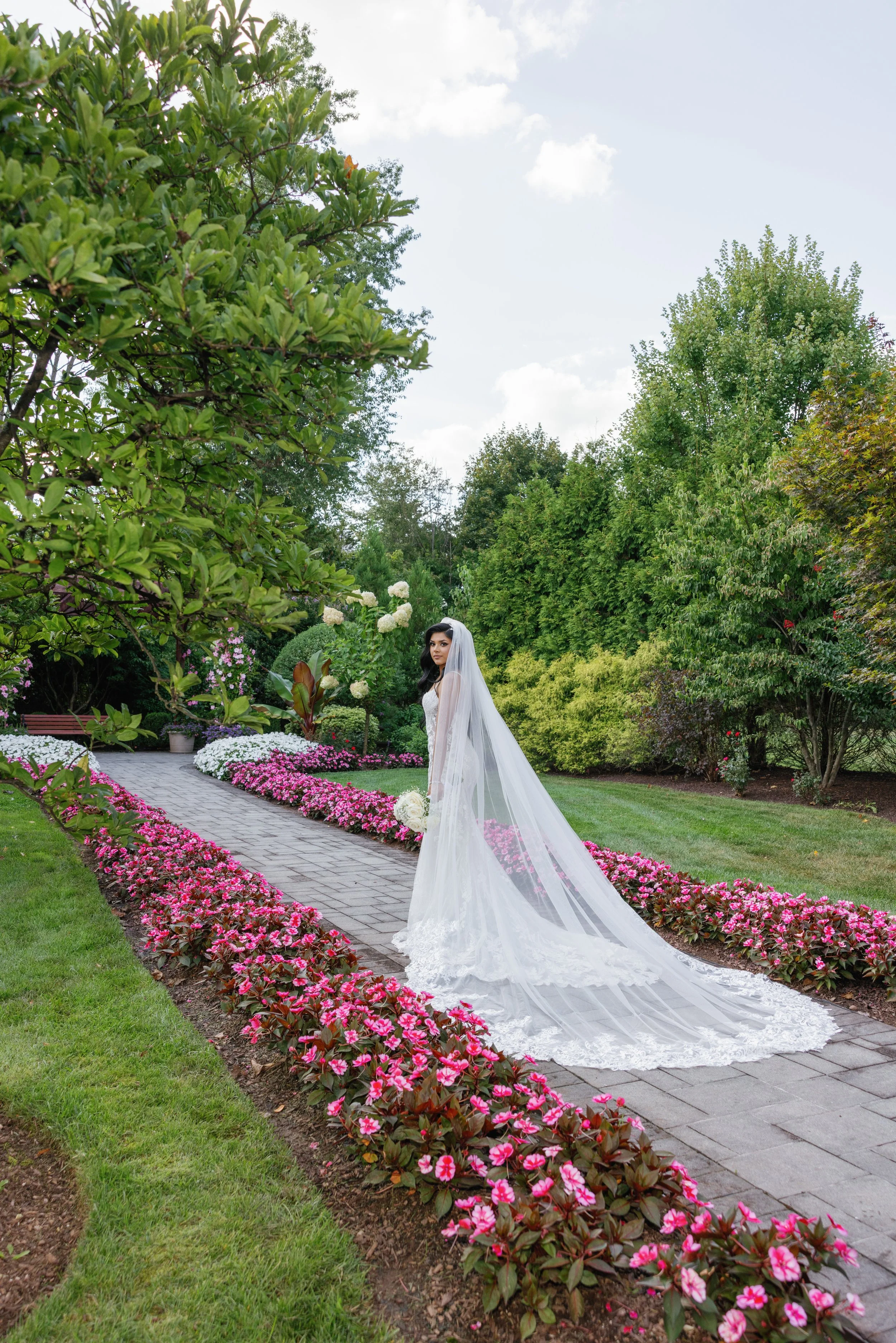 Bride in a white wedding gown with a long veil standing on a garden pathway, surrounded by pink and white flowers and lush green trees.