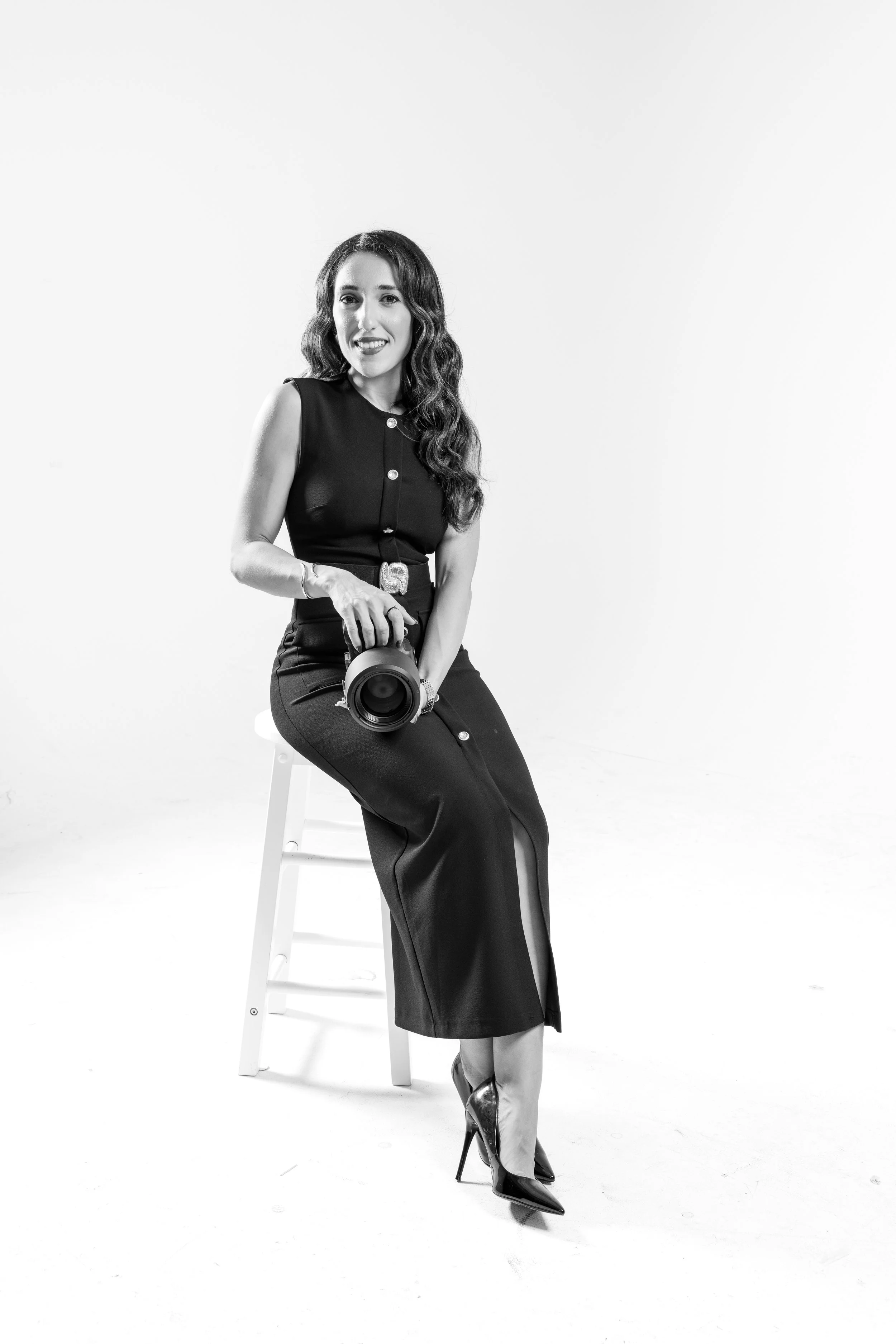 Black and white photo of a woman sitting on a stool, holding a professional camera, wearing a sleeveless top, long skirt with slit, and high heels, smiling at the camera.