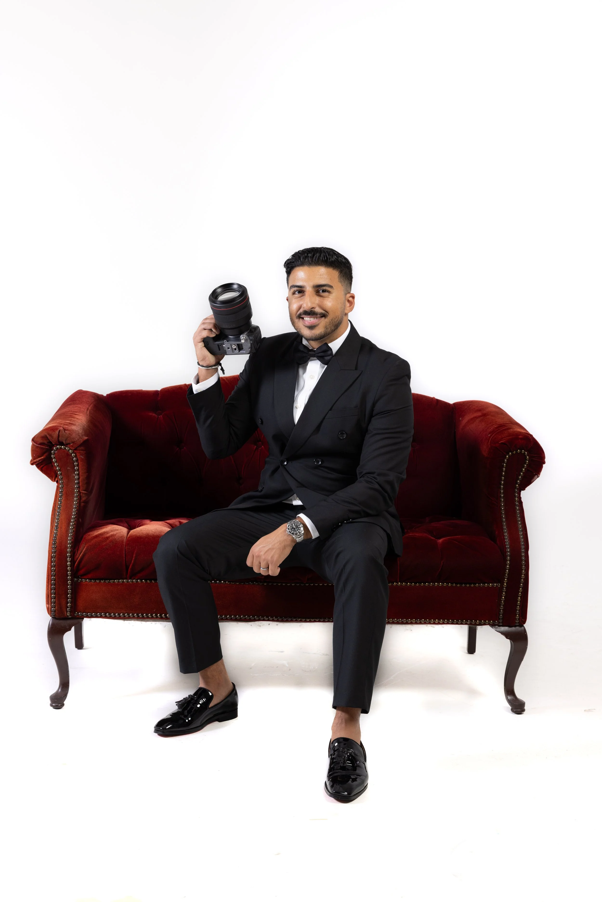 Man in a tuxedo sitting on a red velvet sofa, holding a camera, smiling, against a white background.