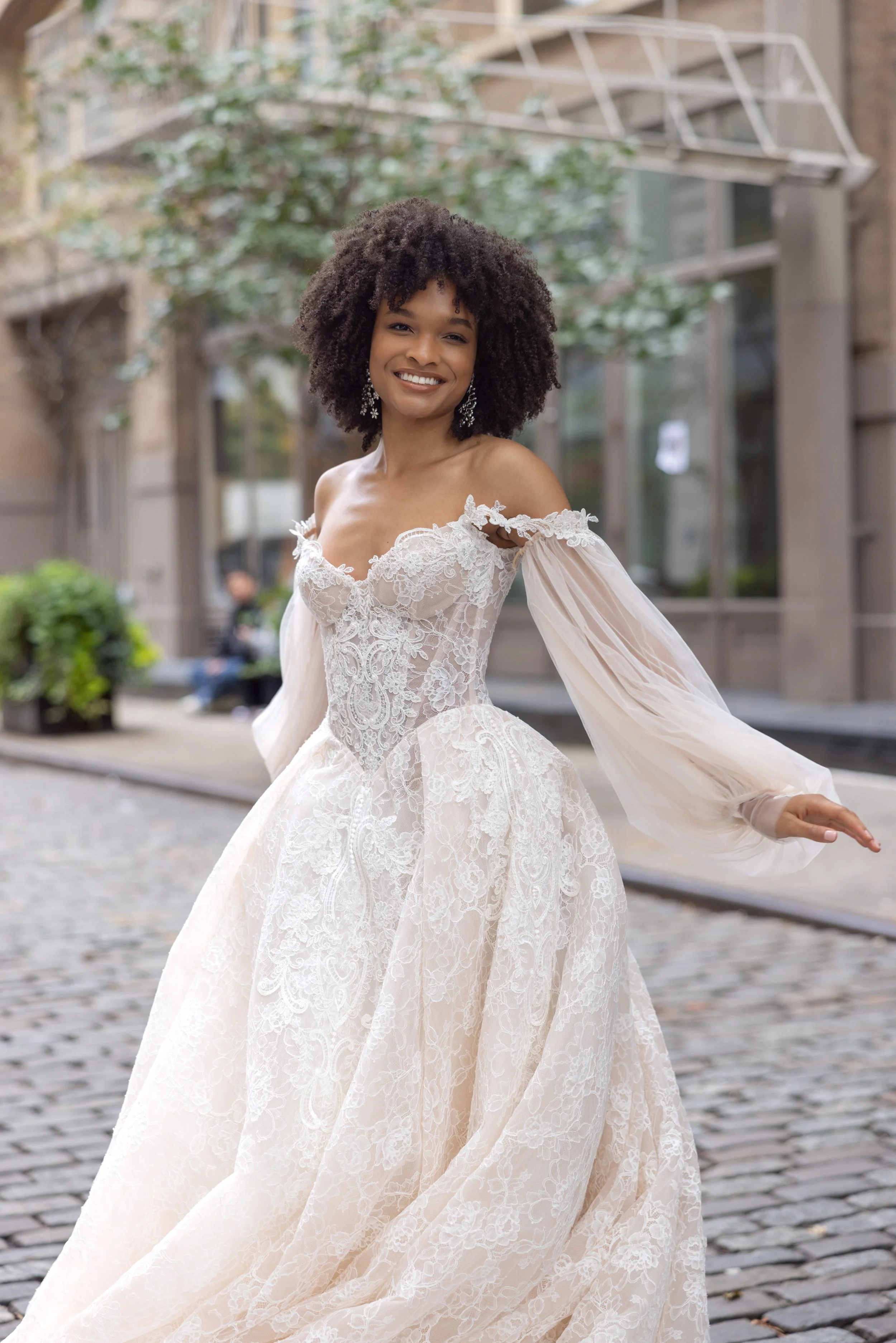 A woman smiling in a wedding dress walking on a city street with cobblestone ground, trees, and buildings in the background.
