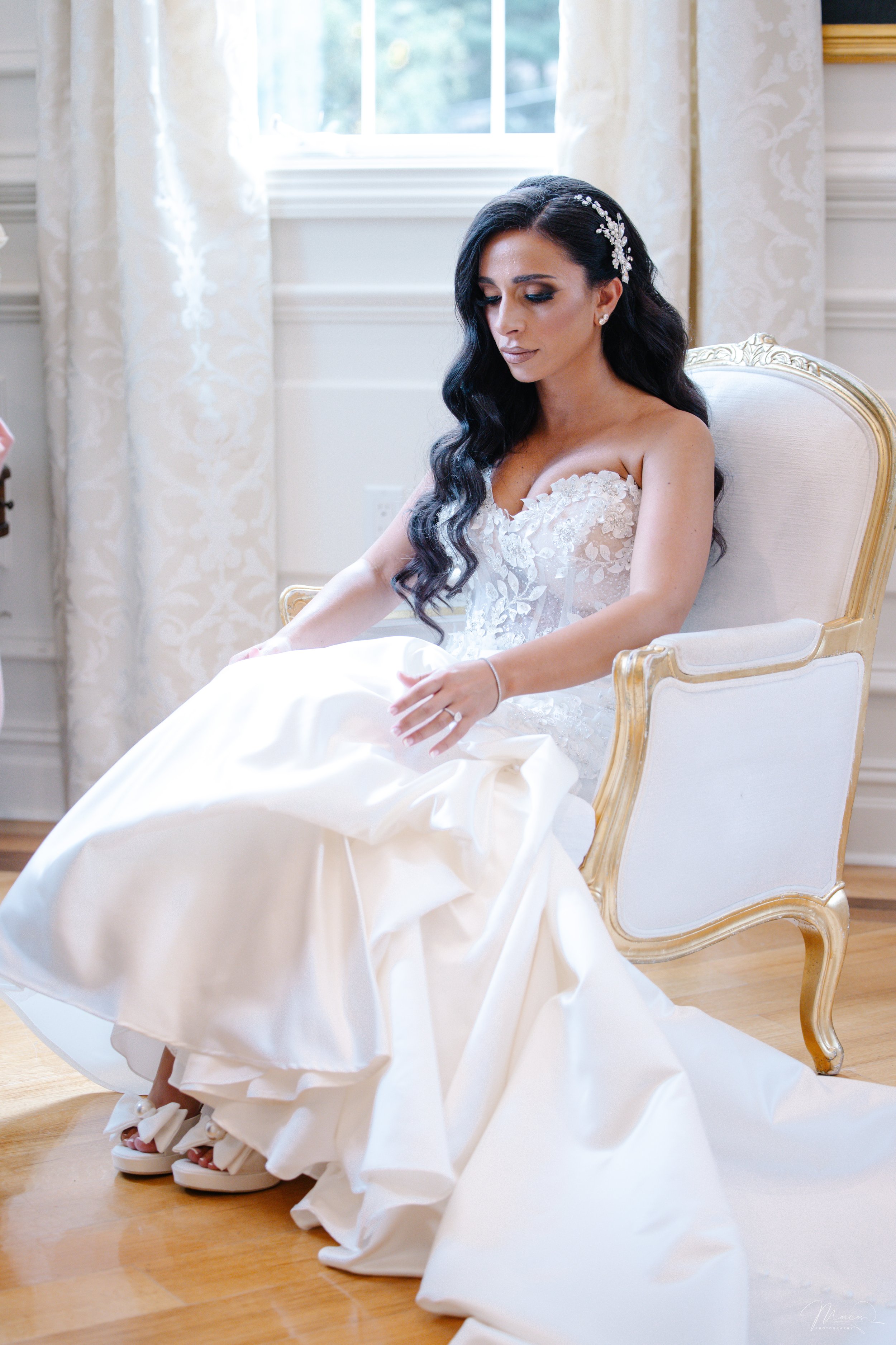 A bride with long dark wavy hair sitting on a white and gold antique chair, wearing a strapless white wedding gown with lace details, adjusting her shoe in a well-lit room with cream curtains and wooden flooring.