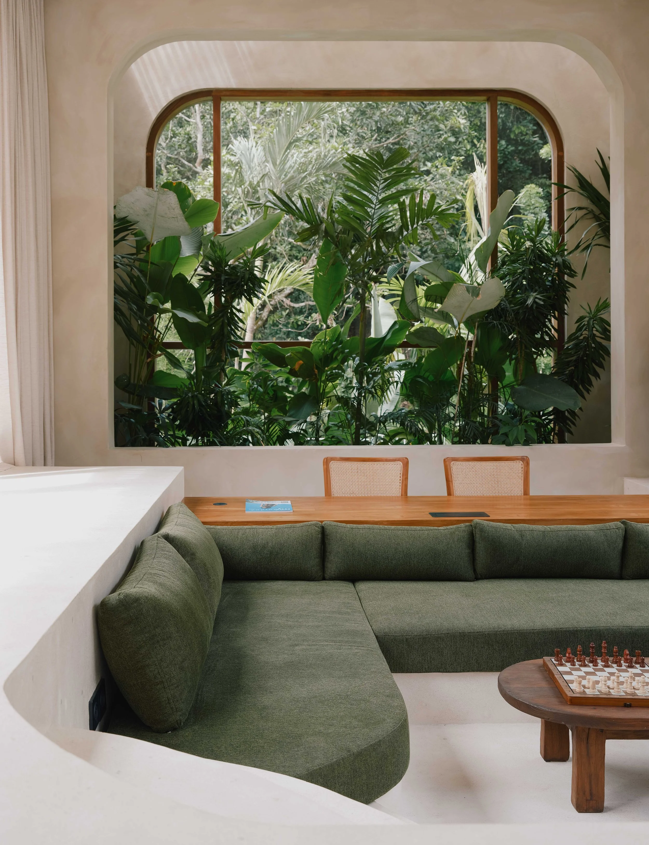 Living room with large window, full of lush green plants, a green sectional sofa, a wooden side table with a chessboard, and a white curved wall.