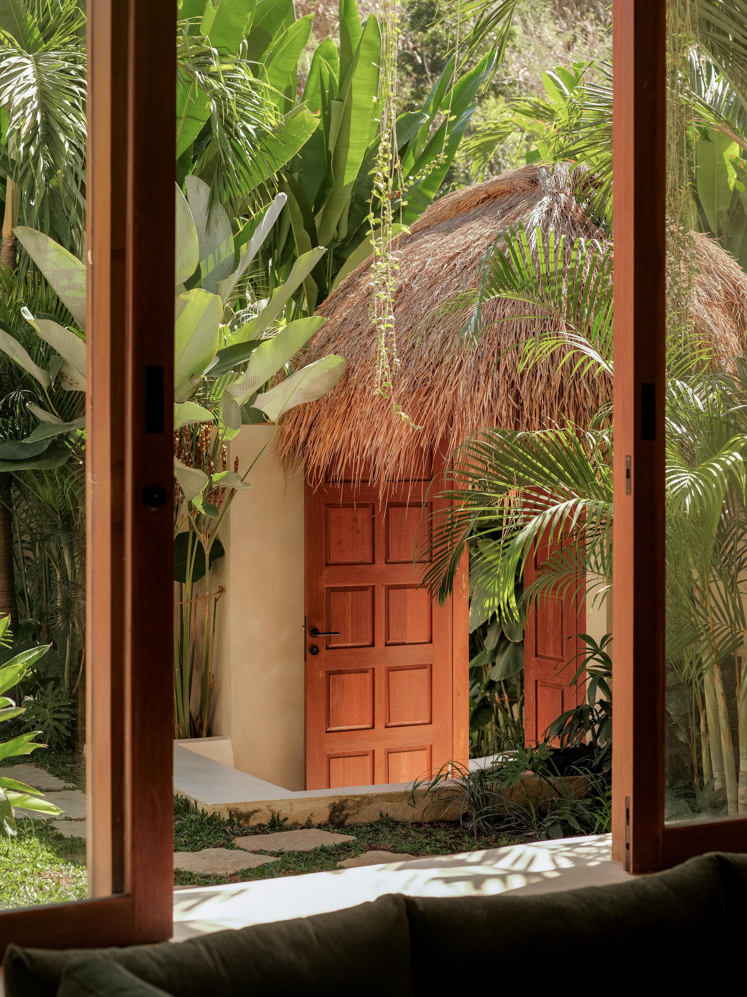View through a window showing tropical plants, a thatched roof structure, and a wooden door, with a stone pathway and lush greenery outside.