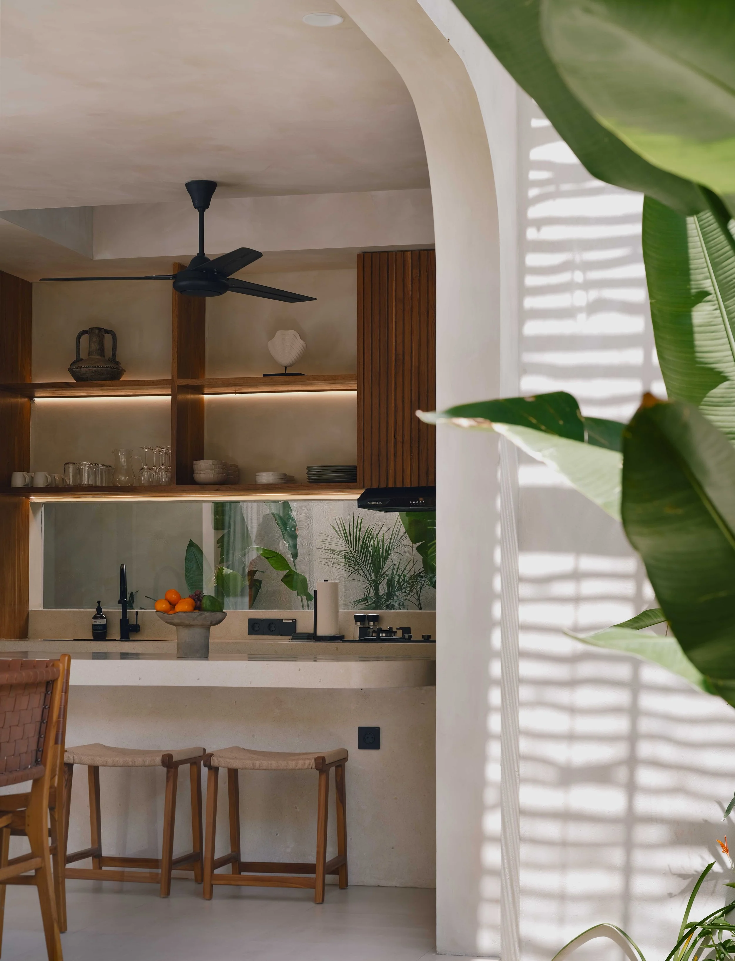 A modern kitchen with a bar counter, three wooden bar stools, a bowl of fresh oranges and limes, a sink, a black faucet, a small plant, wooden shelves with glassware and dishes, a black range hood, a ceiling fan, and ceiling spotlights. There are large green plants and sunlight creating shadows on white curtains.