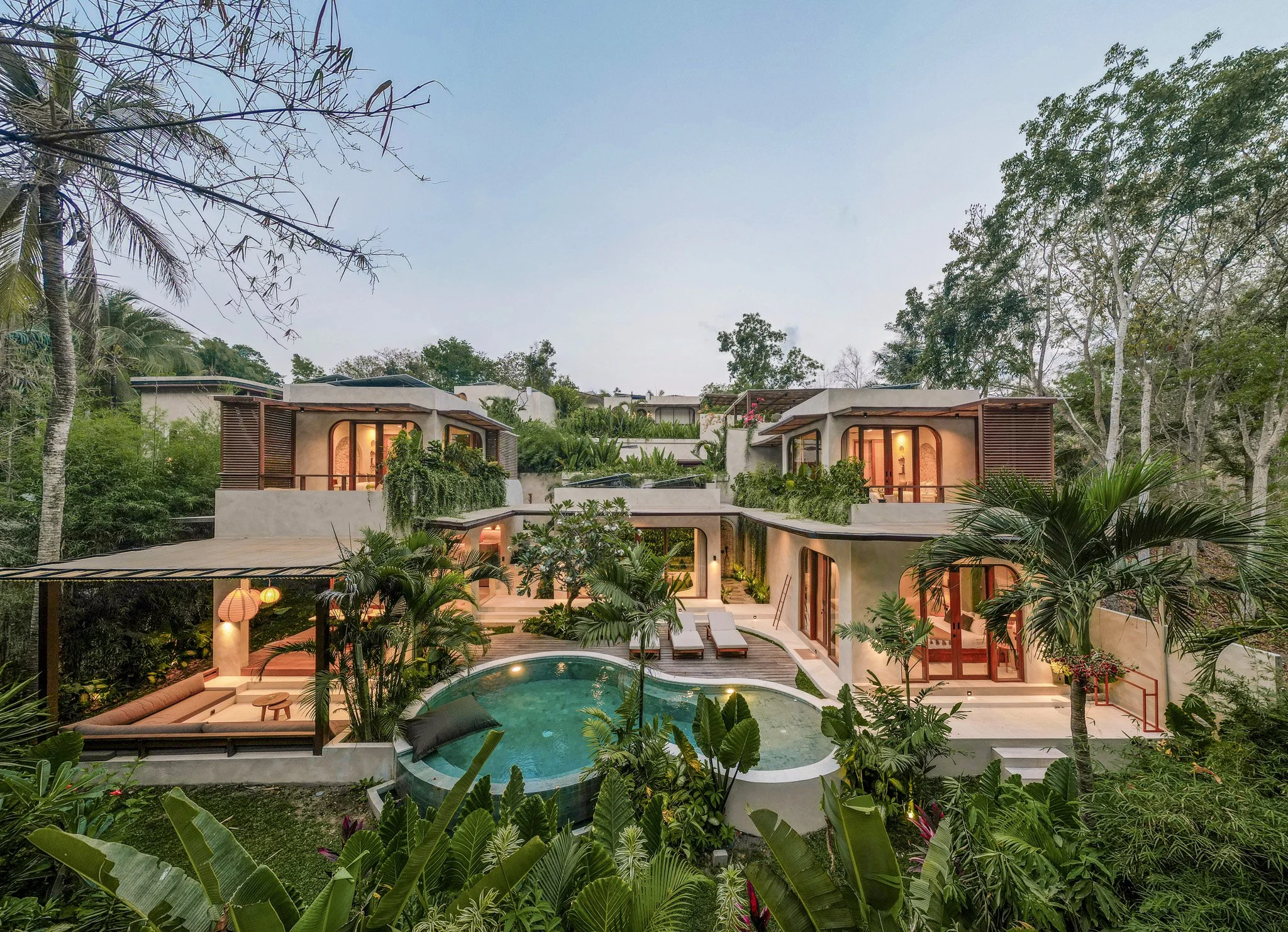 A large, stylish tropical villa with large windows, surrounded by lush green trees and palm trees, with a curved swimming pool and outdoor lounge area.