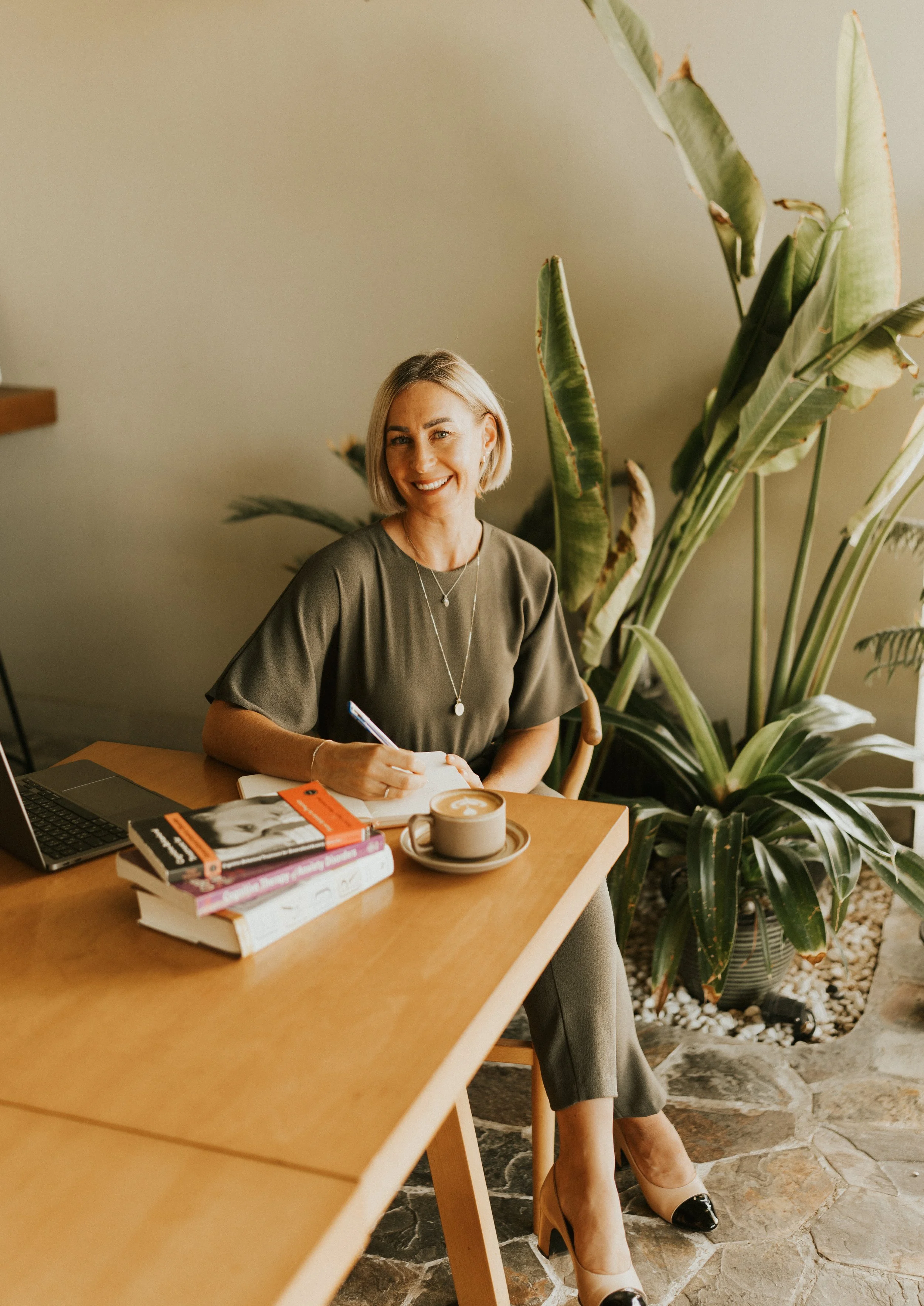 Kat Cuthbert EMDR & CBT Therapist BABCP Accredited, smiling, seated at a wooden table with a laptop, notebooks, and a cup of coffee, in a room with large green plants and stone flooring.