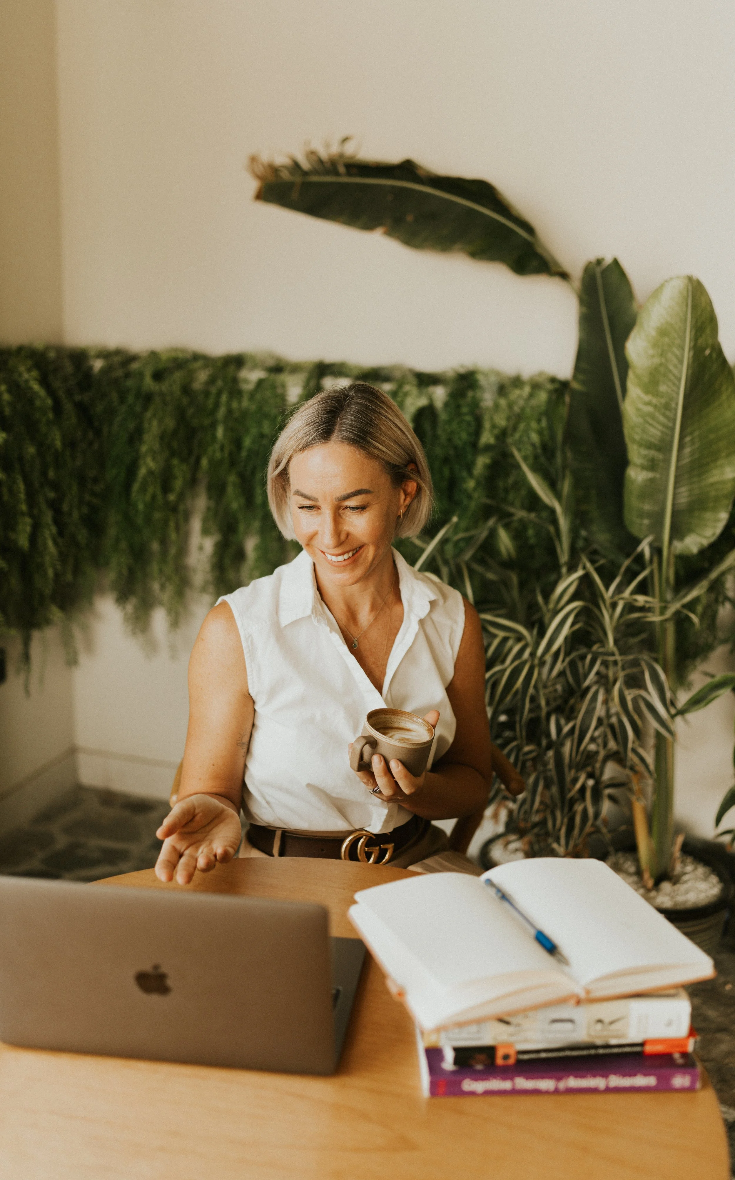 Kat Cuthbert EMDR & CBT Therapist BABCP Accredited sitting at a wooden table with an open laptop and stacked books, holding a cup of coffee or tea, smiling in front of a green plant wall.
