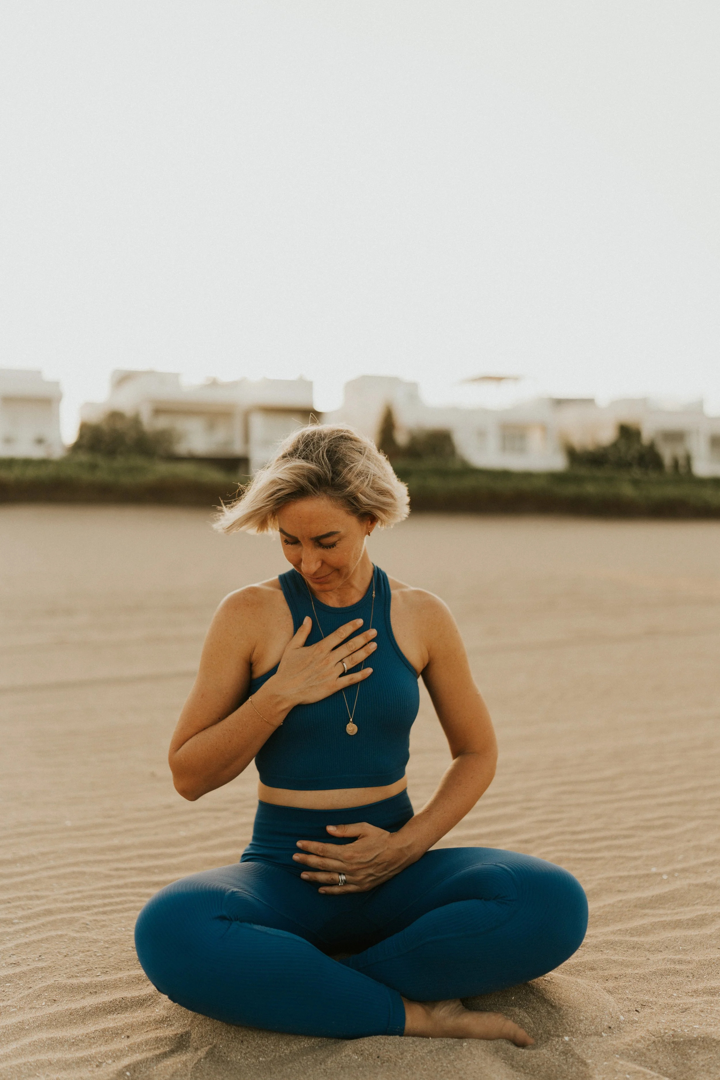 Kat Cuthbert EMDR & CBT Therapist BABCP Accredited practicing yoga on a sandy beach, sitting cross-legged in a blue workout outfit, with one hand on her chest and the other on her stomach.