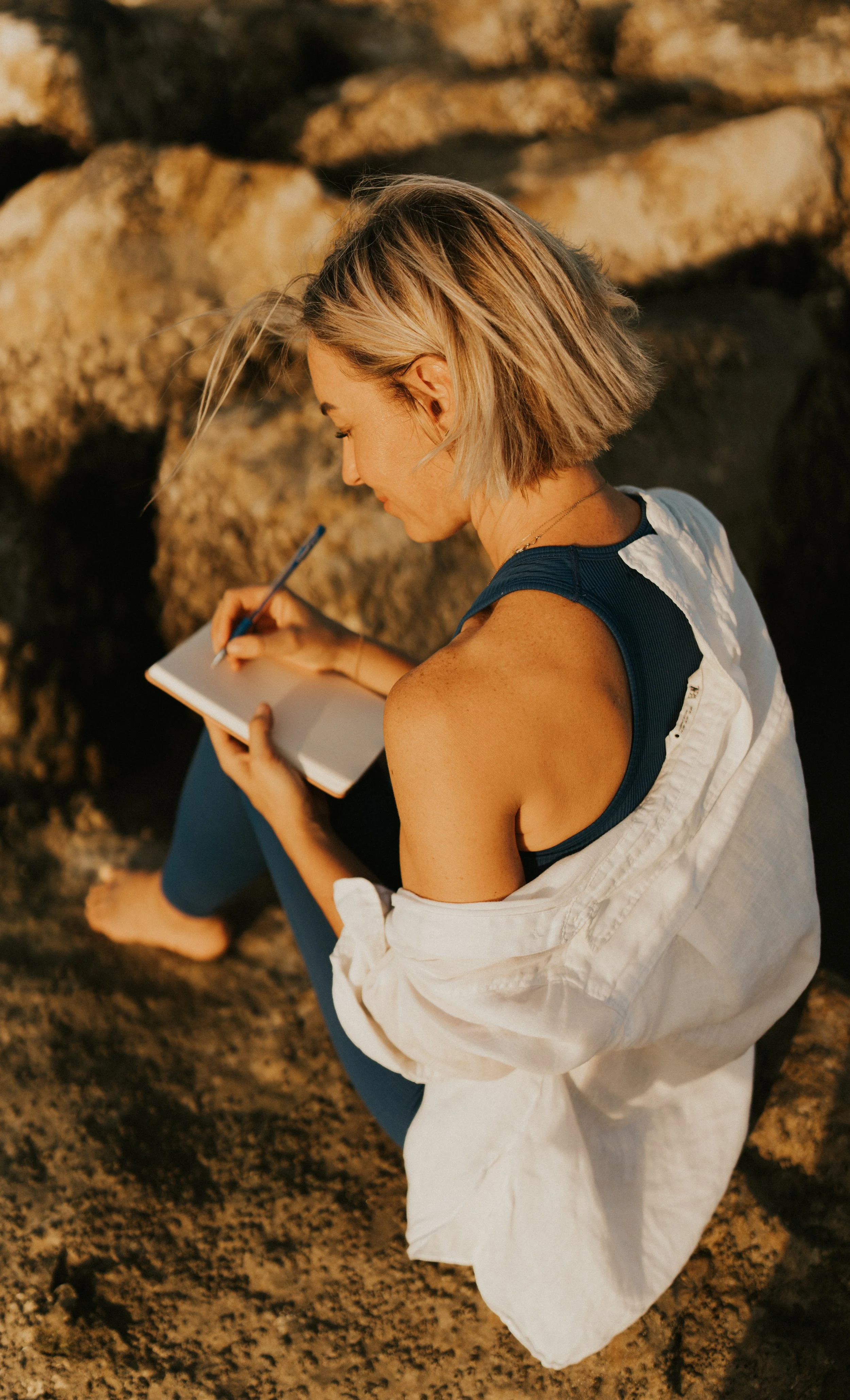Kat Cuthbert EMDR & CBT Therapist BABCP Accredited wearing a sleeveless navy top and a white shirt, sitting on rocks, writing in a notebook with a pen during sunset.