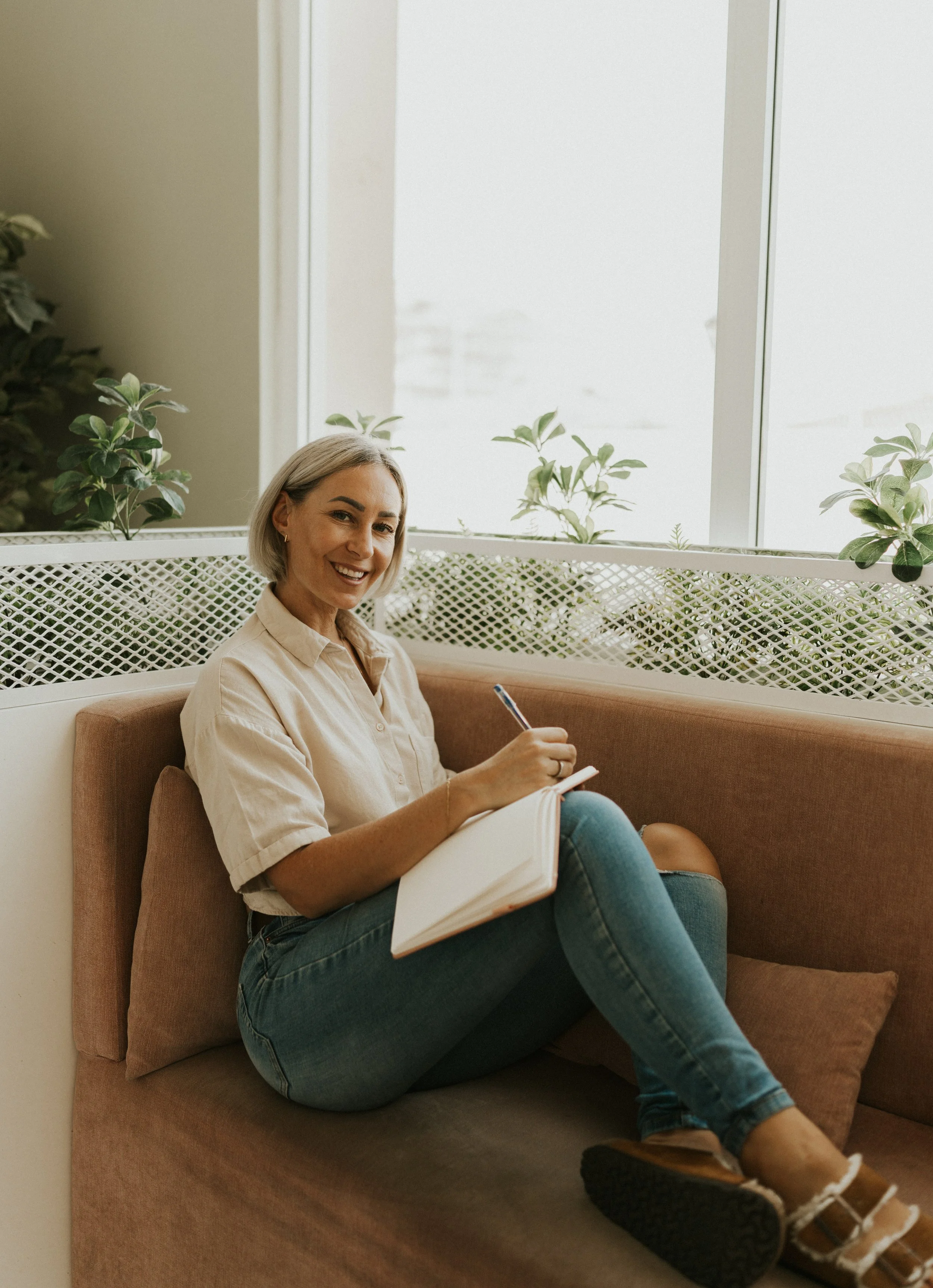 Kat Cuthbert EMDR & CBT Therapist BABCP Accredited sitting on a pink cushioned bench, smiling, holding a pen, and writing in a notebook. There are green plants and a large window behind her.