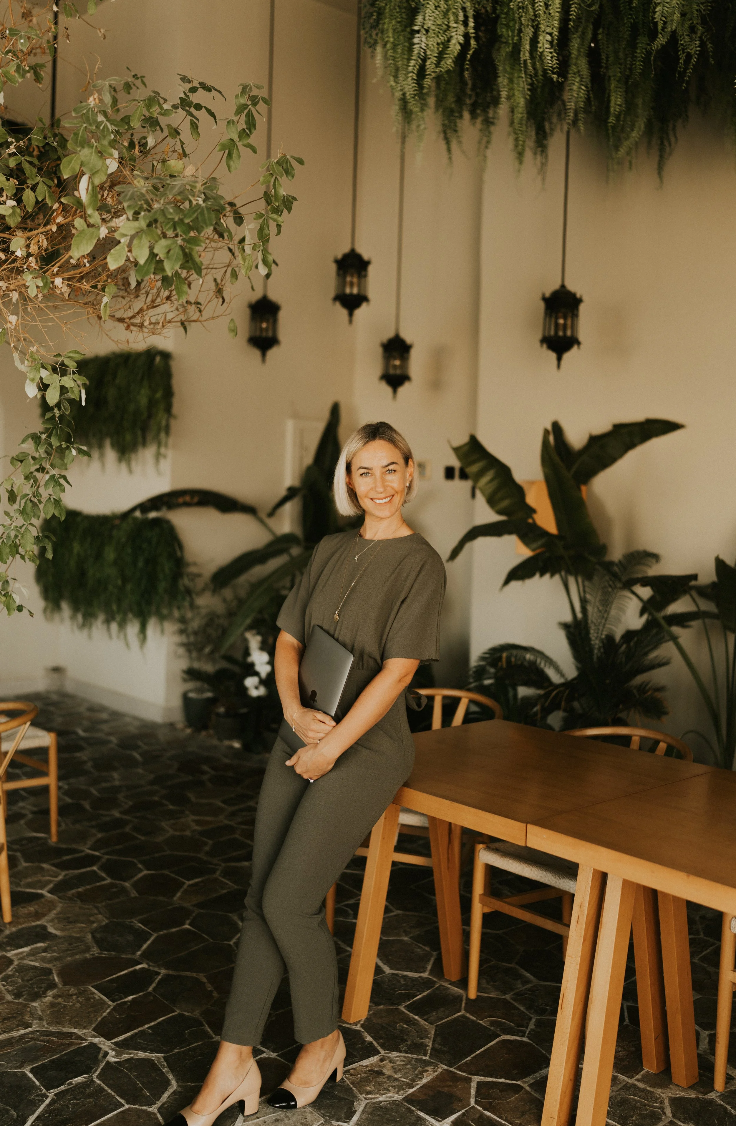 Kat Cuthbert EMDR & CBT Therapist BABCP Accredited with a brown outfit holding a closed laptop, standing in a modern room with hanging lanterns and lush green plants.