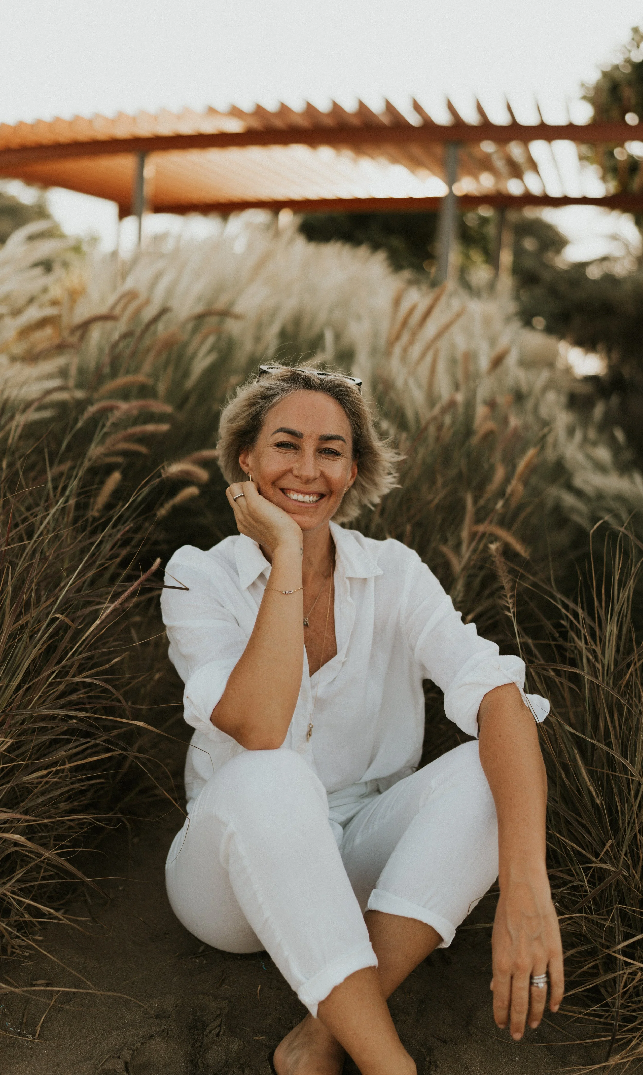 Kat Cuthbert EMDR & CBT Therapist BABCP Accredited wearing a white shirt and white pants, sitting cross-legged on the ground in a field of tall grasses, smiling at the camera.