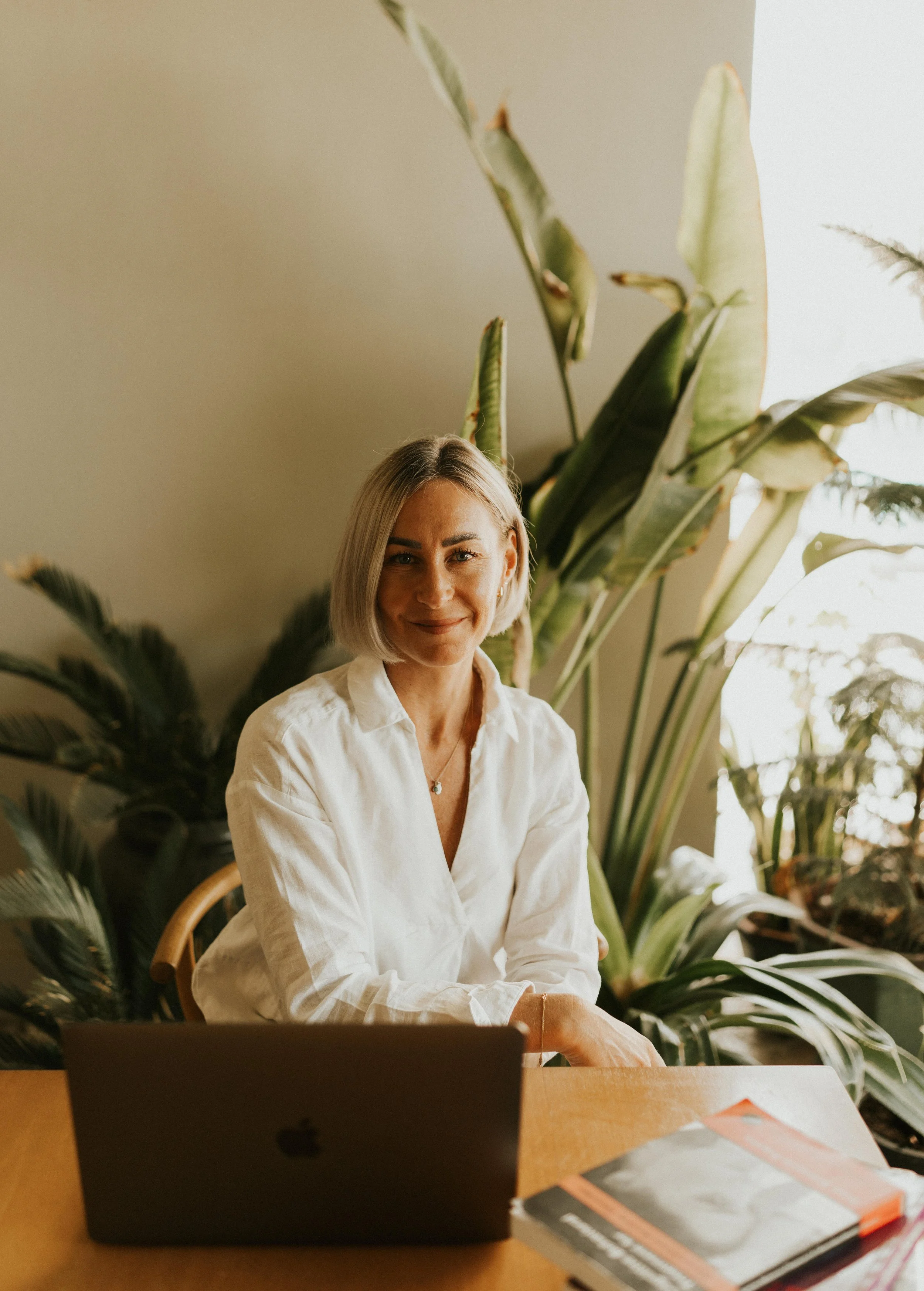 Kat Cuthbert EMDR & CBT Therapist BABCP Accredited wearing  white blouse sitting at a wooden table in front of a laptop, with green plants behind her.