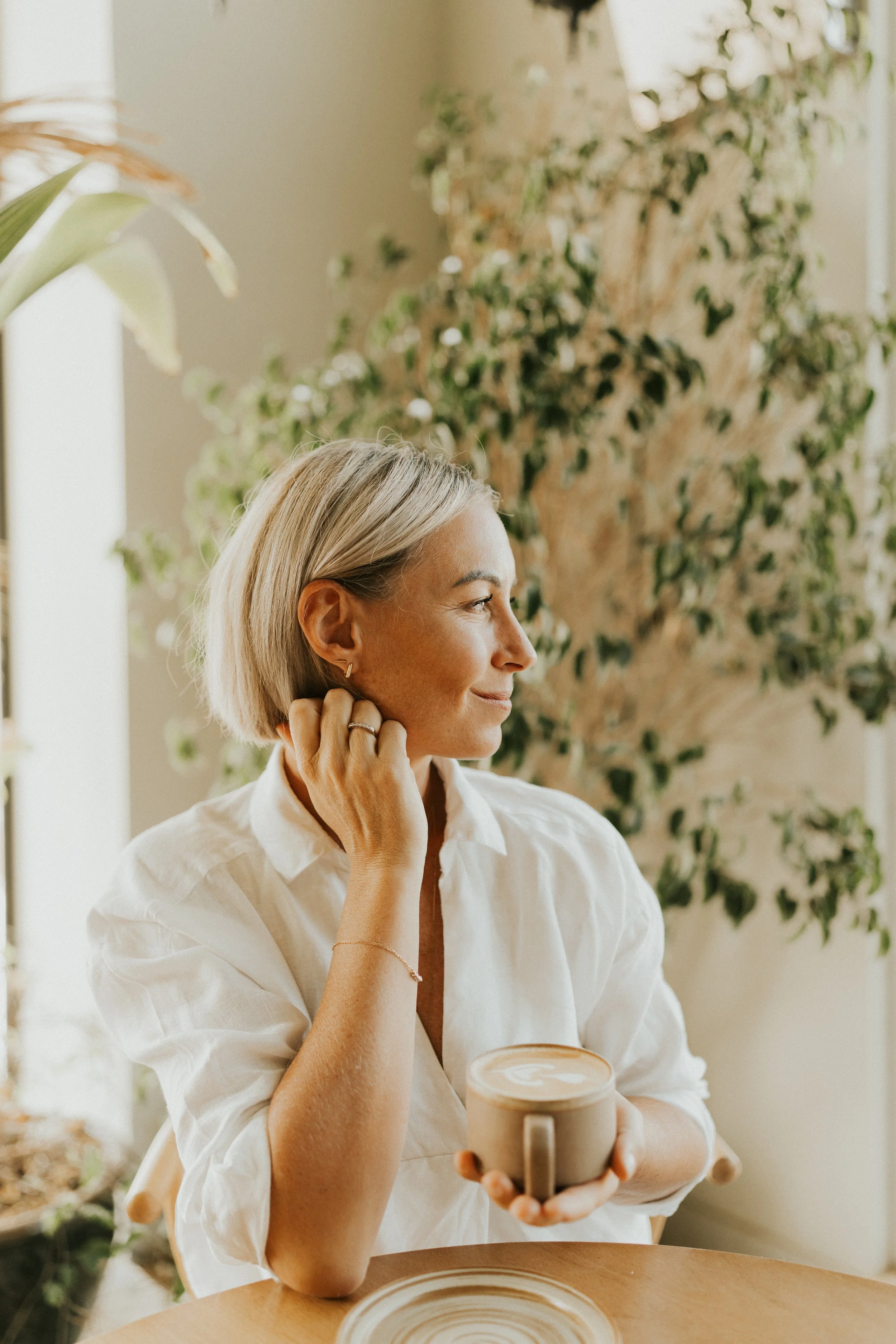 Kat Cuthbert EMDR & CBT Therapist BABCP Accredited wearing a white shirt, holding a coffee mug, sitting at a wooden table with plants in the background.
