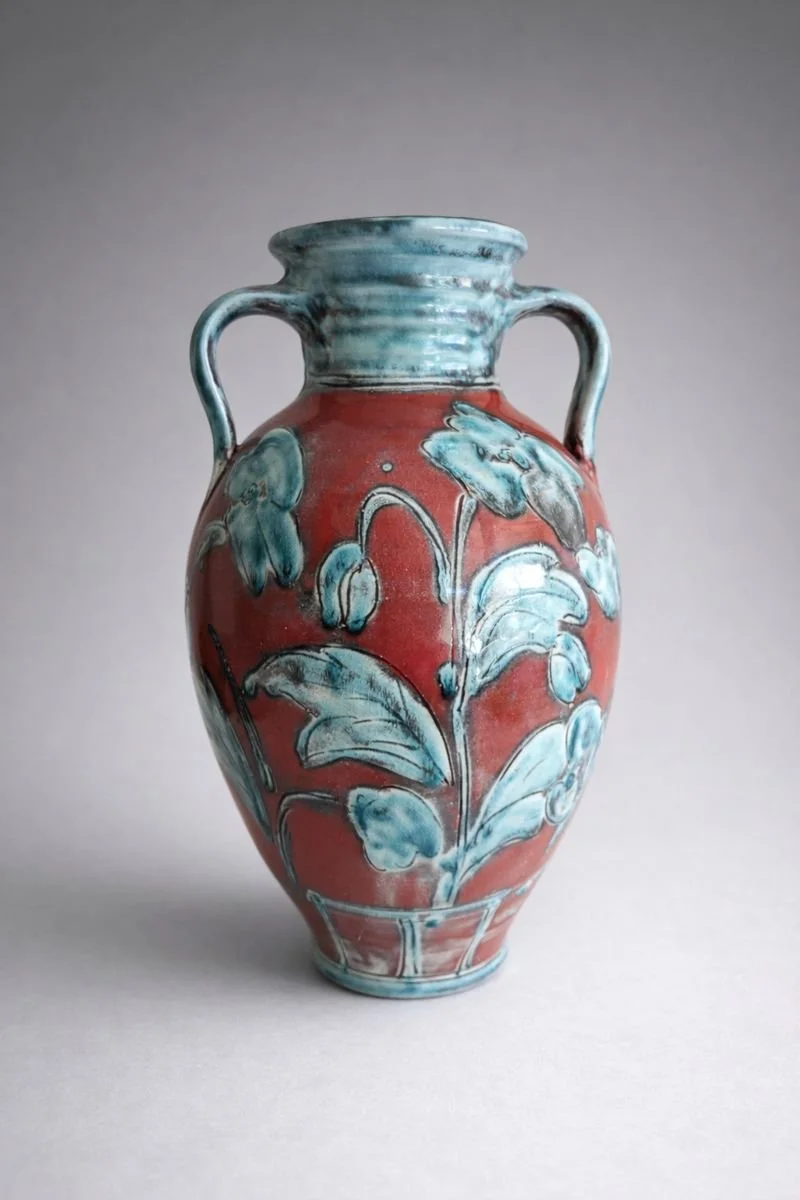 A ceramic vase with a red background and blue floral patterns, featuring two handles and a rounded body.
