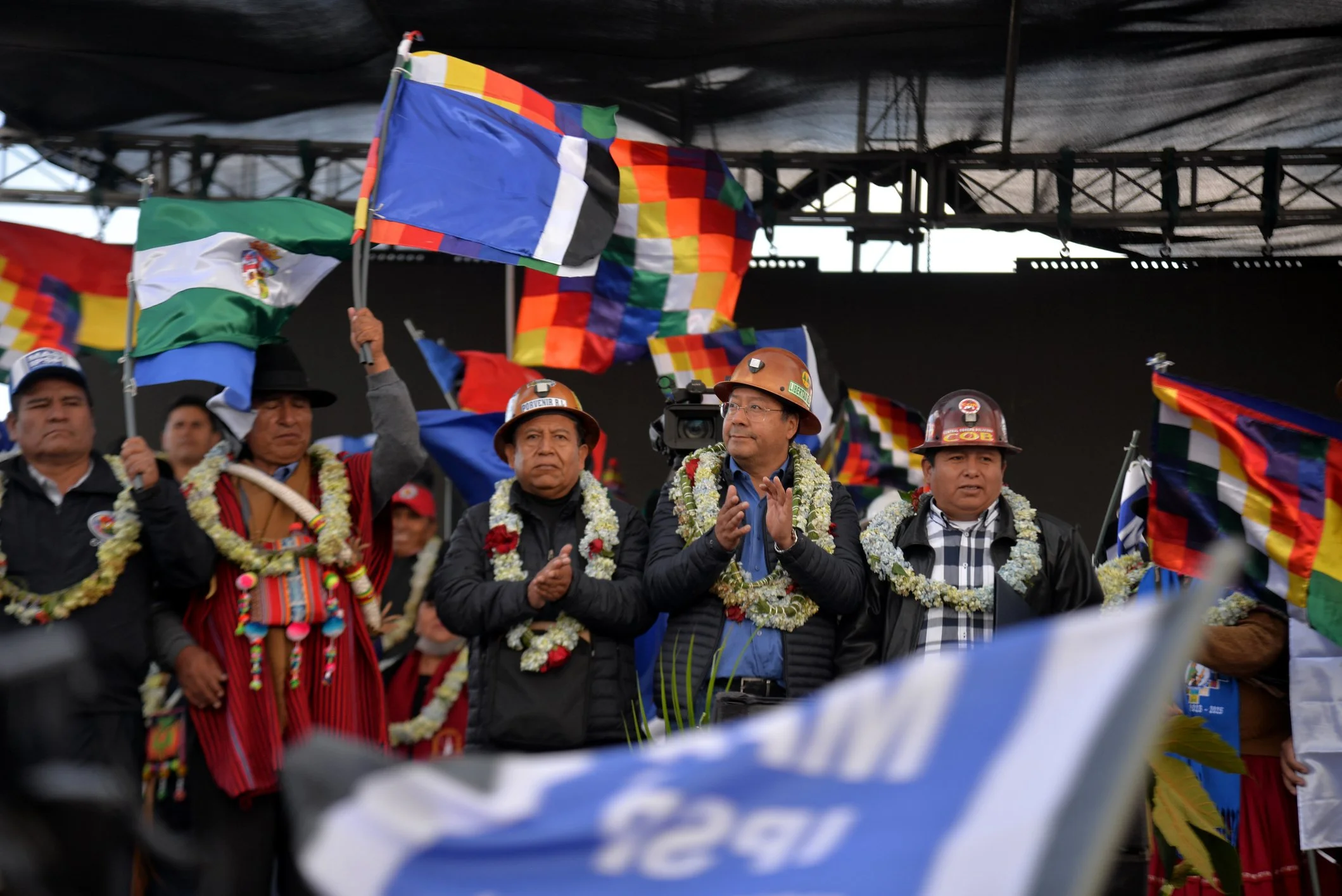Un grupo de personas en una ceremonia, usando guirnaldas y cascos, con banderas de colores brillante y ropa tradicional en un escenario cubierto