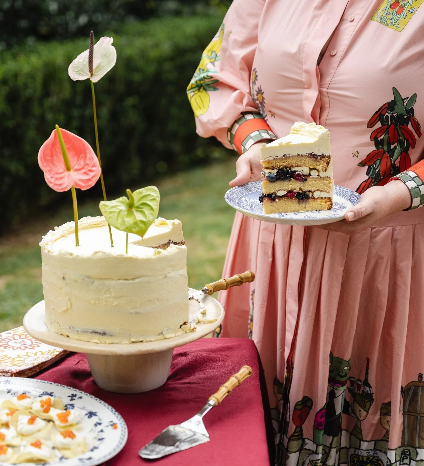 Person holding a plate with a slice of layered berry cake at a celebration with a decorated cake on a table in an outdoor setting.