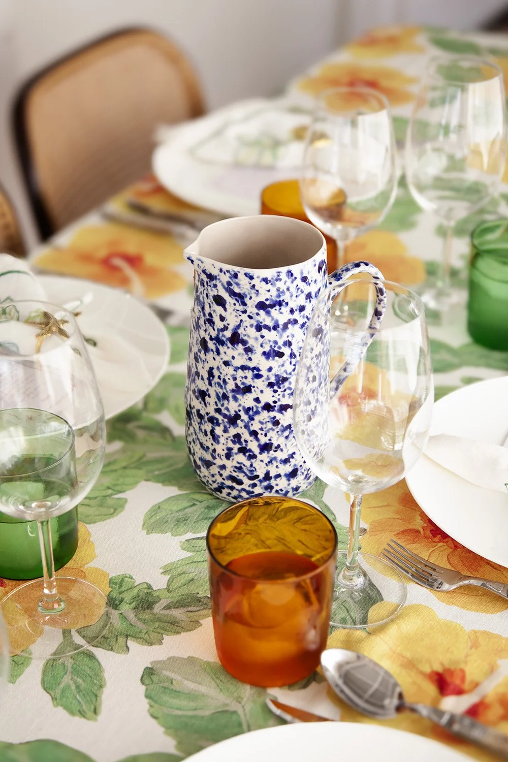 A table set for a meal with a colorful floral tablecloth, a blue and white speckled ceramic pitcher, various colored glasses, white plates, and silverware.