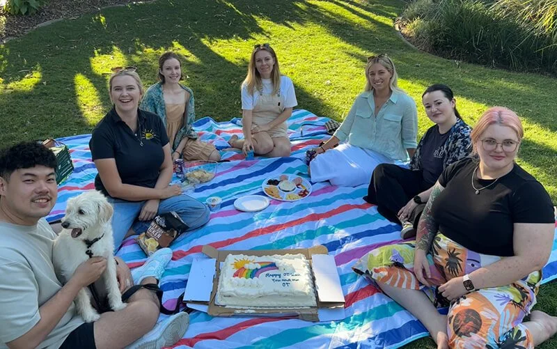 Group of seven people and a dog sitting on a picnic blanket outdoors, with a birthday cake and snacks on a table, enjoying a sunny day.