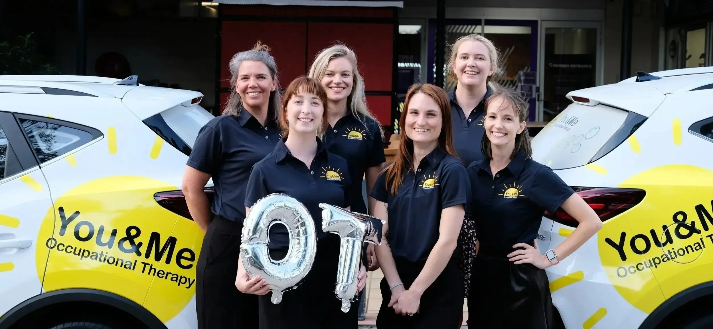 Group of six women standing in front of two cars with yellow and black branding, holding silver balloons forming the number 01, all wearing matching navy blue polo shirts with a yellow sun logo and the text "You & Me Occupational Therapy."
