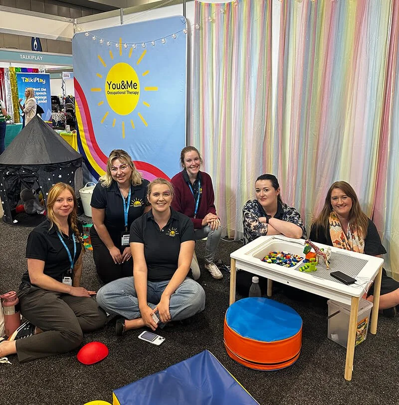 Group of six women sitting and kneeling in front of a colorful display booth for You & Me Occupational Therapy at an event or expo, with toys and therapy tools on a small white table.