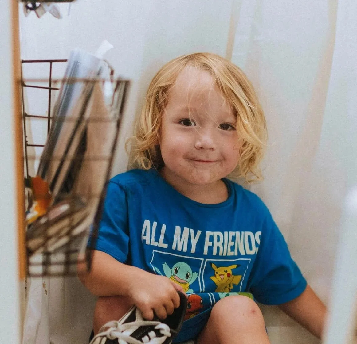 Young boy with curly blonde hair sitting on the floor in a corner, wearing a blue Pokémon T-shirt, smiling at the camera, holding a shoe in his hand.