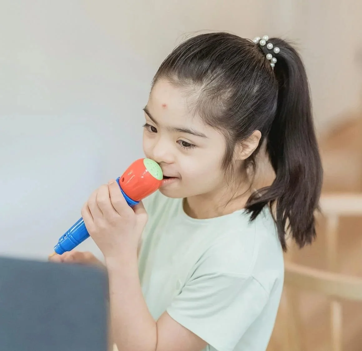 A young girl with a ponytail holding a toy microphone and looking at a laptop screen.