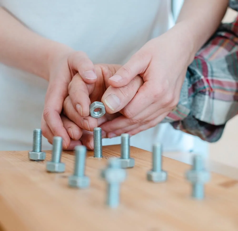 Person holding a hex nut above a wooden surface with several metal bolts scattered on it.