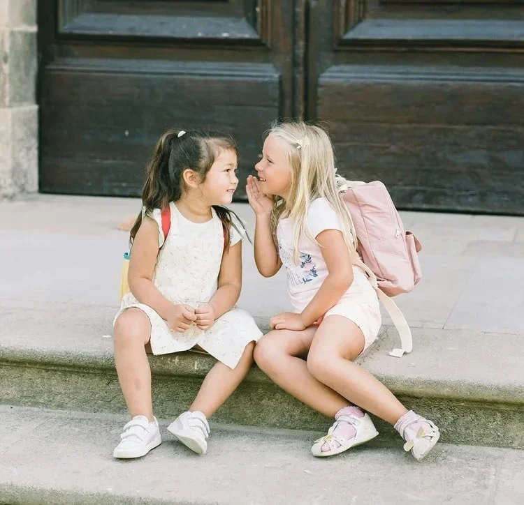 Two young girls sitting on concrete steps in front of a wooden door, engaged in a secret conversation, with backpacks on their shoulders.