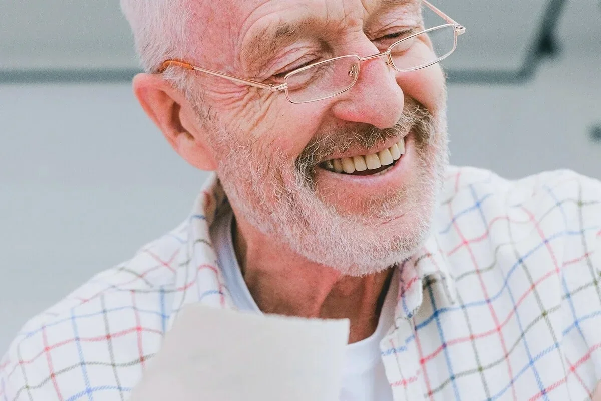 Close-up of an elderly man smiling happily, wearing glasses and a checkered shirt.