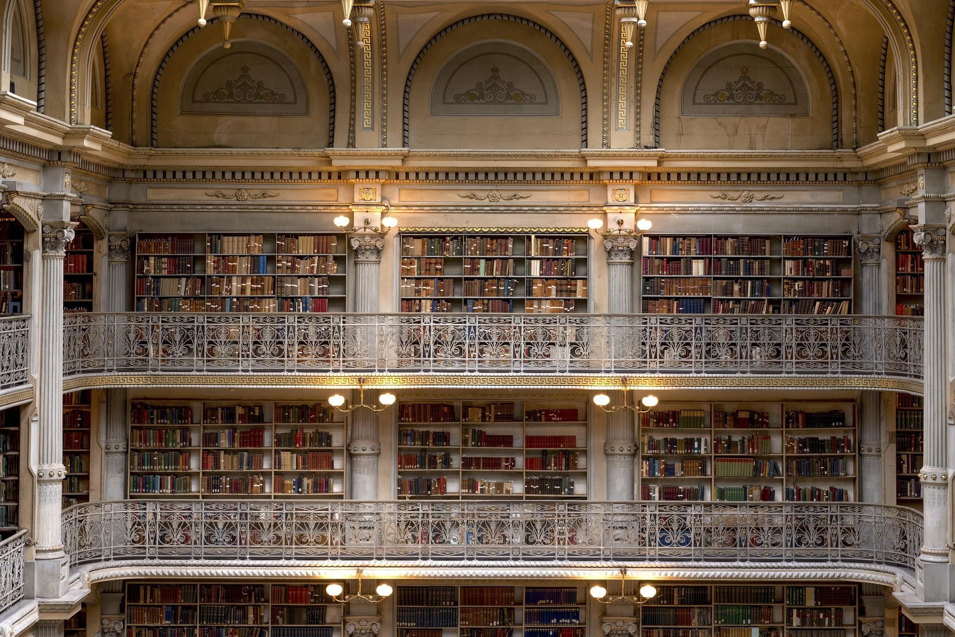 Interior view of a multi-level historic library with ornate railings, columns, and bookshelves filled with books, lit by vintage-style lamps.