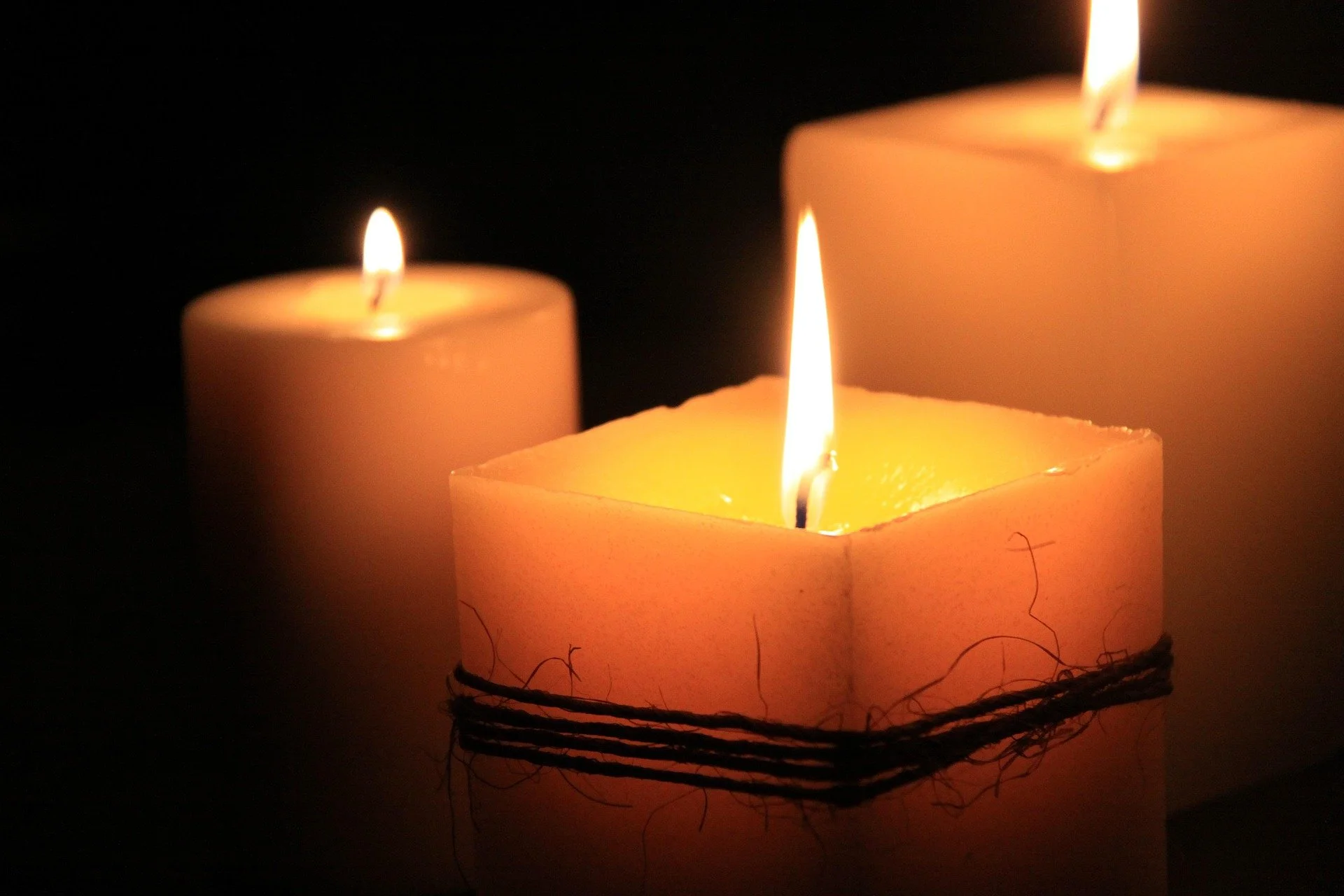 Three lit candles in dark surroundings, two rectangular and one cylindrical, with warm flickering flames.
