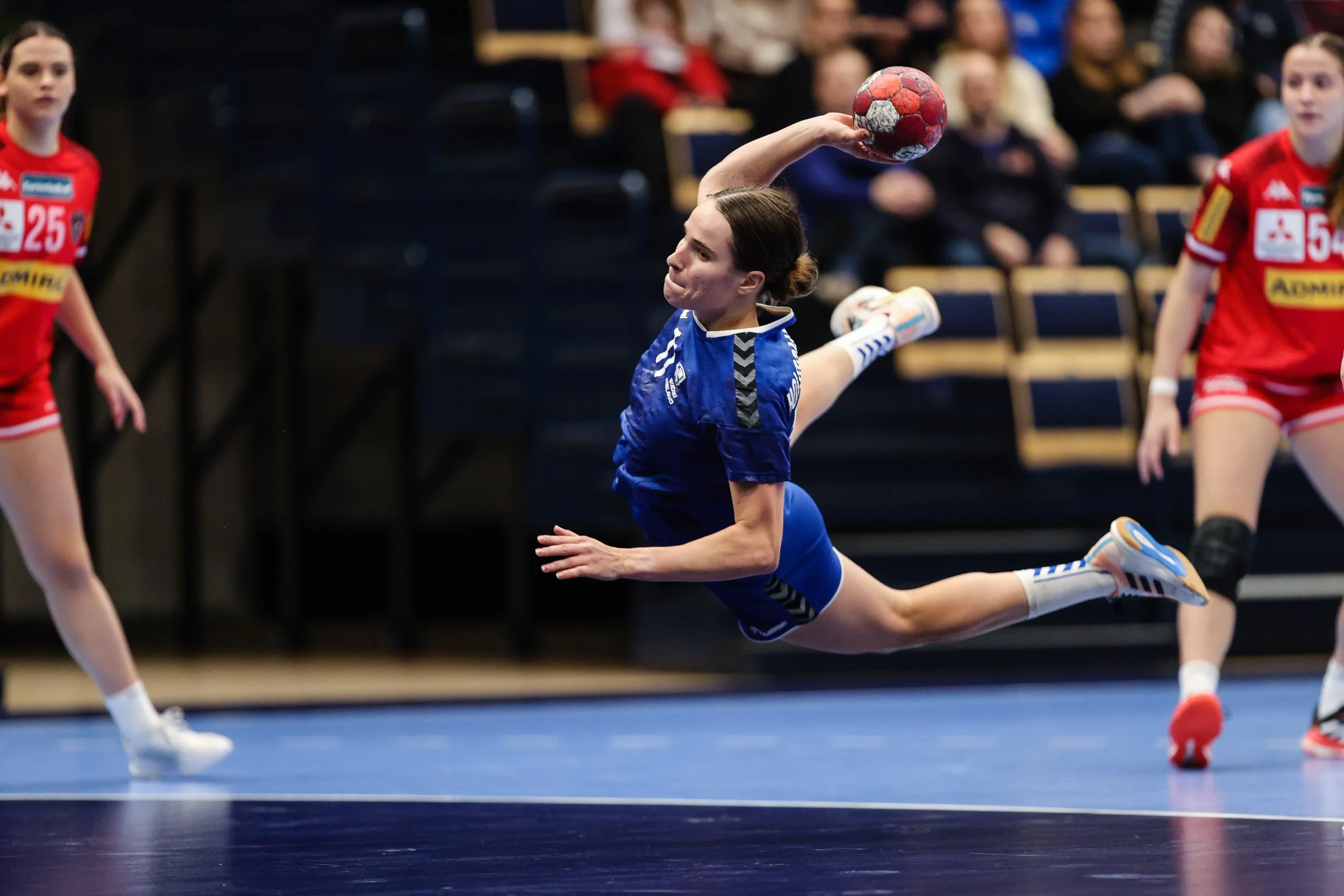 A female athlete in a blue jersey and shorts is in mid-air, diving to throw a handball during a game, with two other players in red jerseys nearby and spectators in the background.