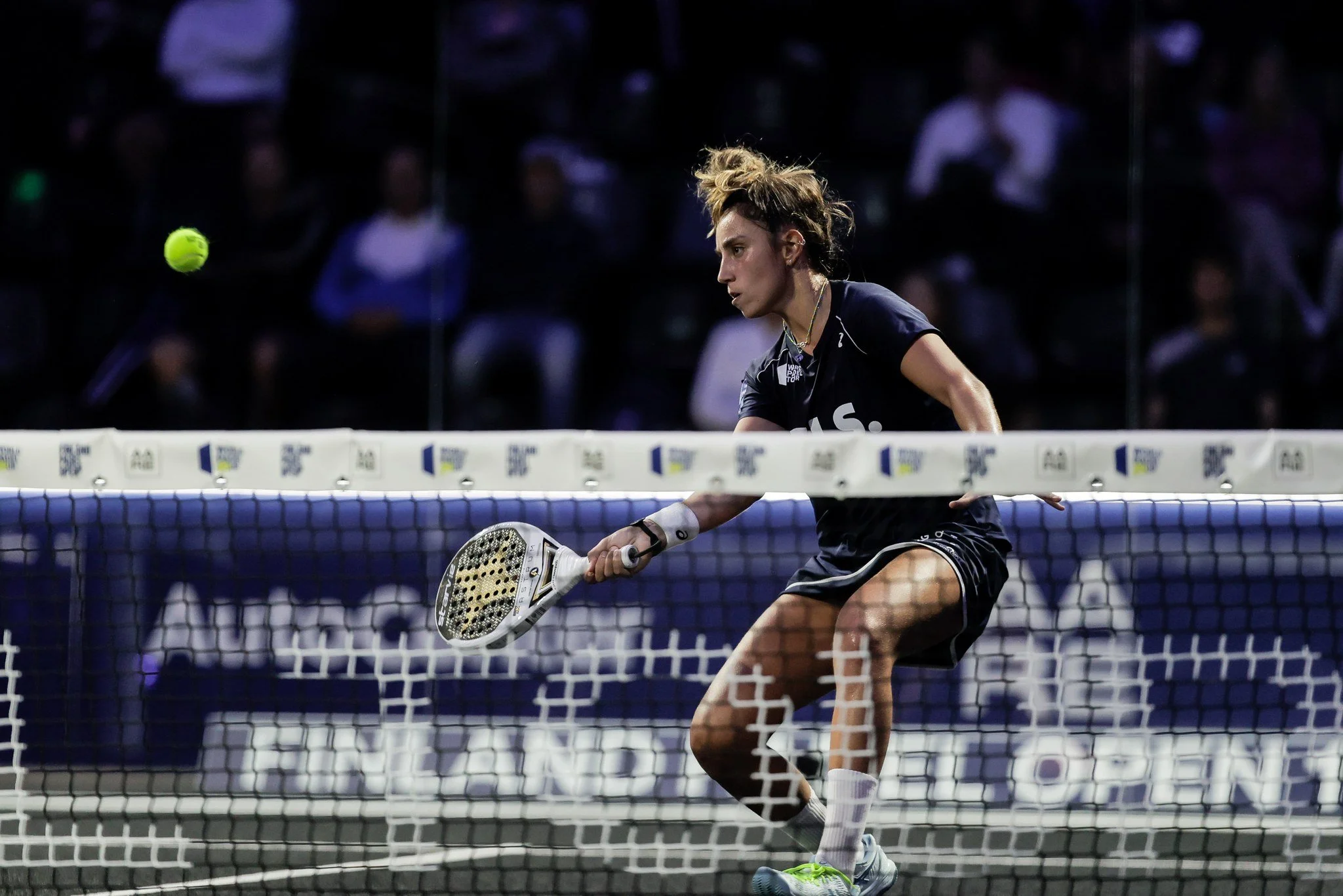 A female padel player in a black uniform playing a match on an indoor court. She is hitting a ball with a racket near the net, with spectators watching in the background.