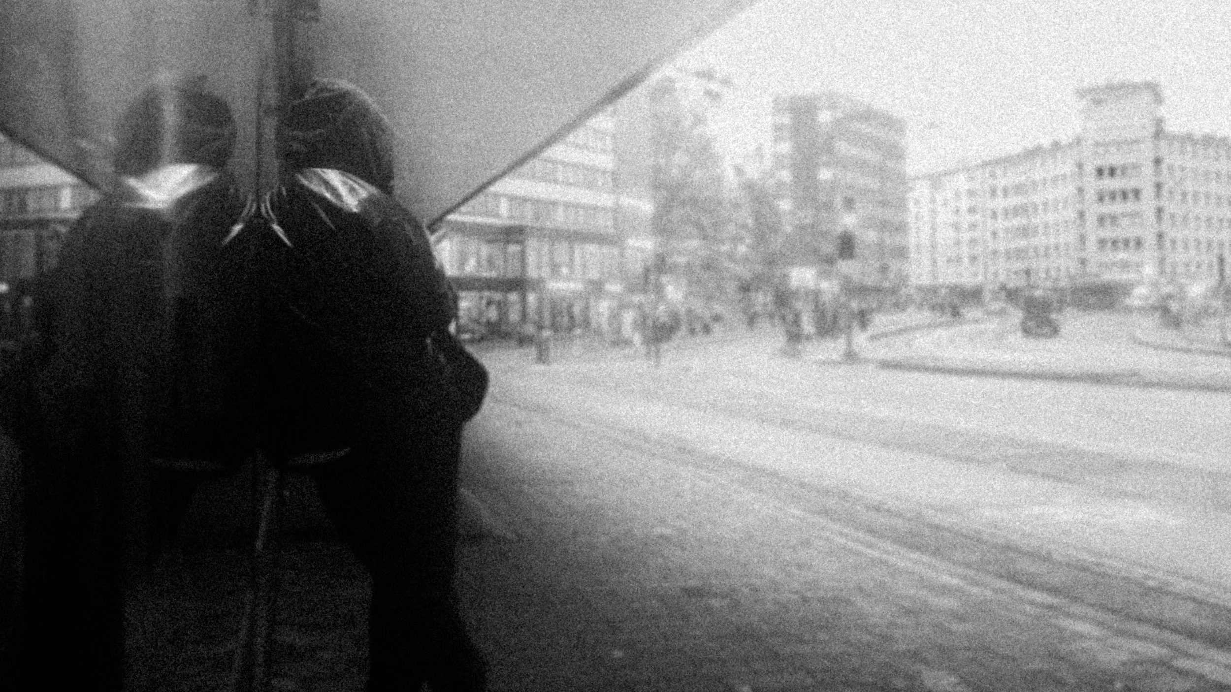 Two people standing on a city sidewalk with tram tracks, buildings, and a busy street in the background, black and white photo.
