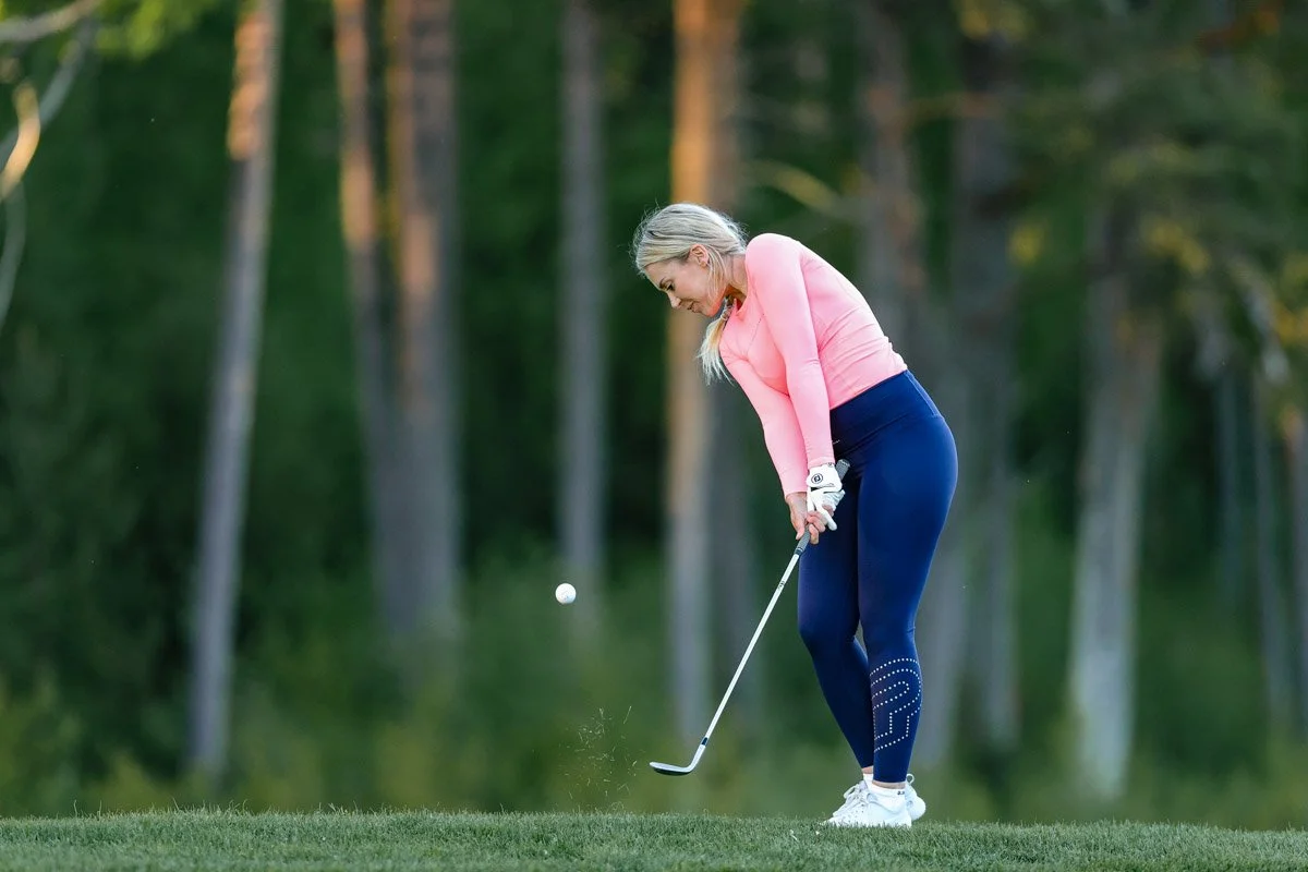 A woman in pink and blue athletic attire playing golf on a course with trees in the background.