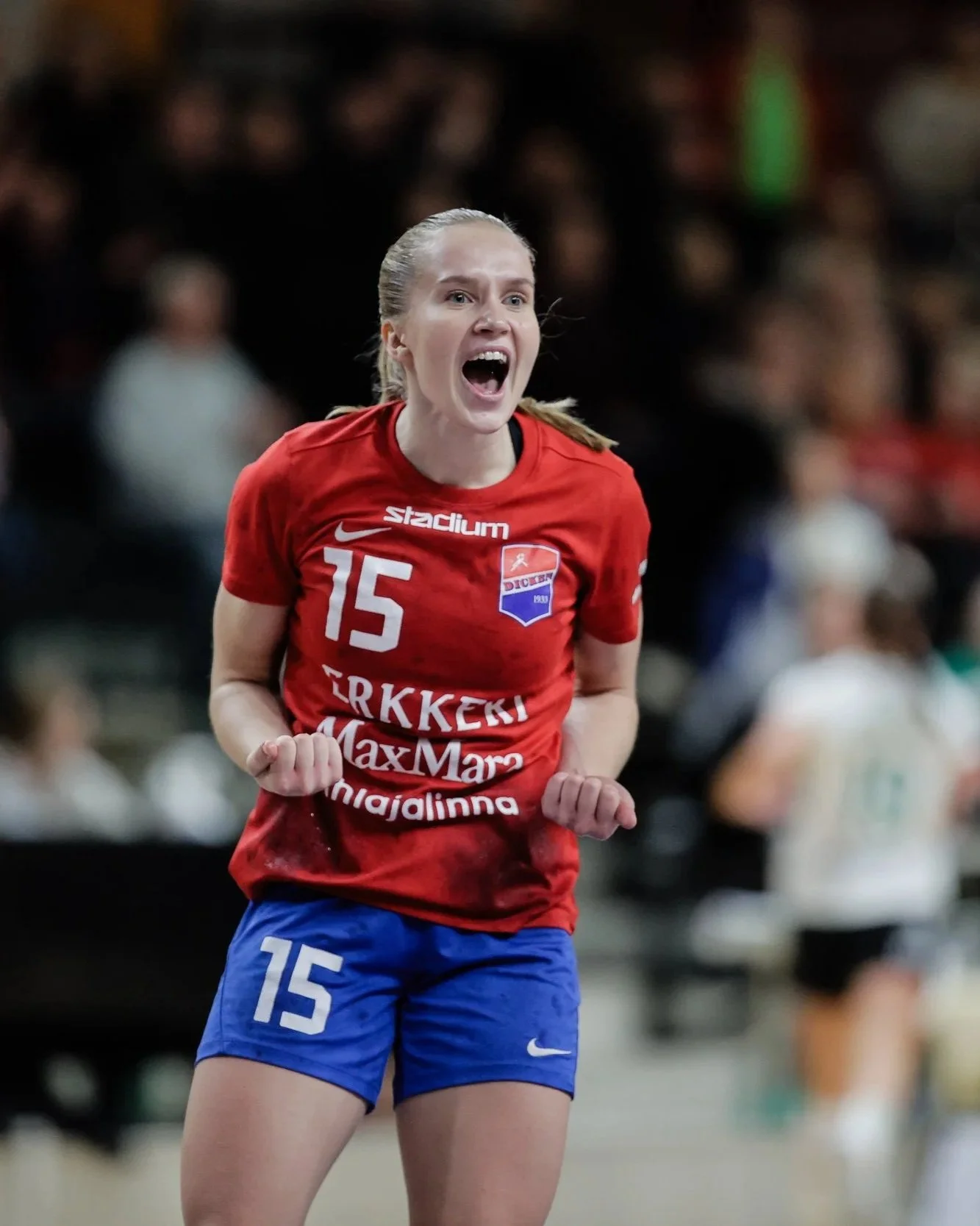 A female handball player celebrating on the court, wearing a red jersey with the number 15 and blue shorts. She appears excited with her fists clenched.