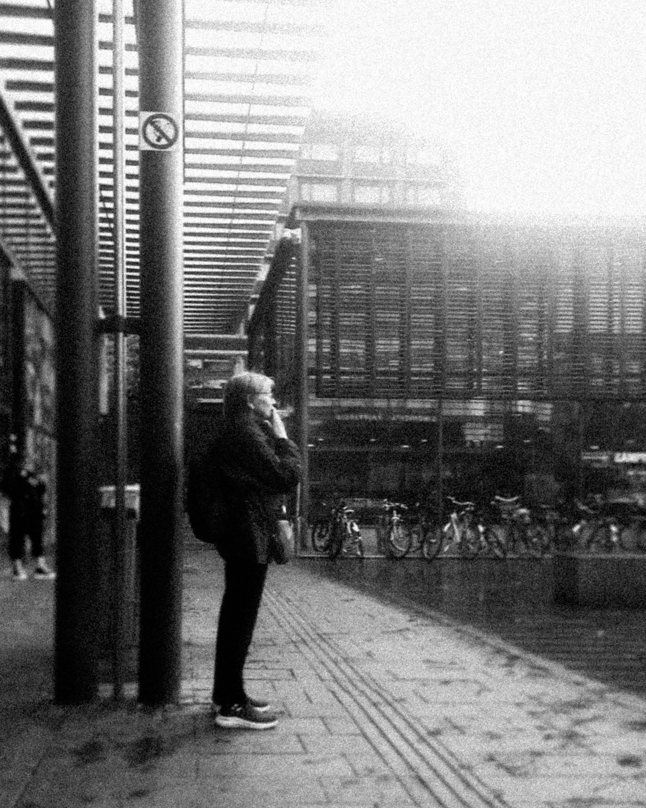 A person standing on a city sidewalk, talking on a cell phone, with bicycles parked in the background and modern buildings around.