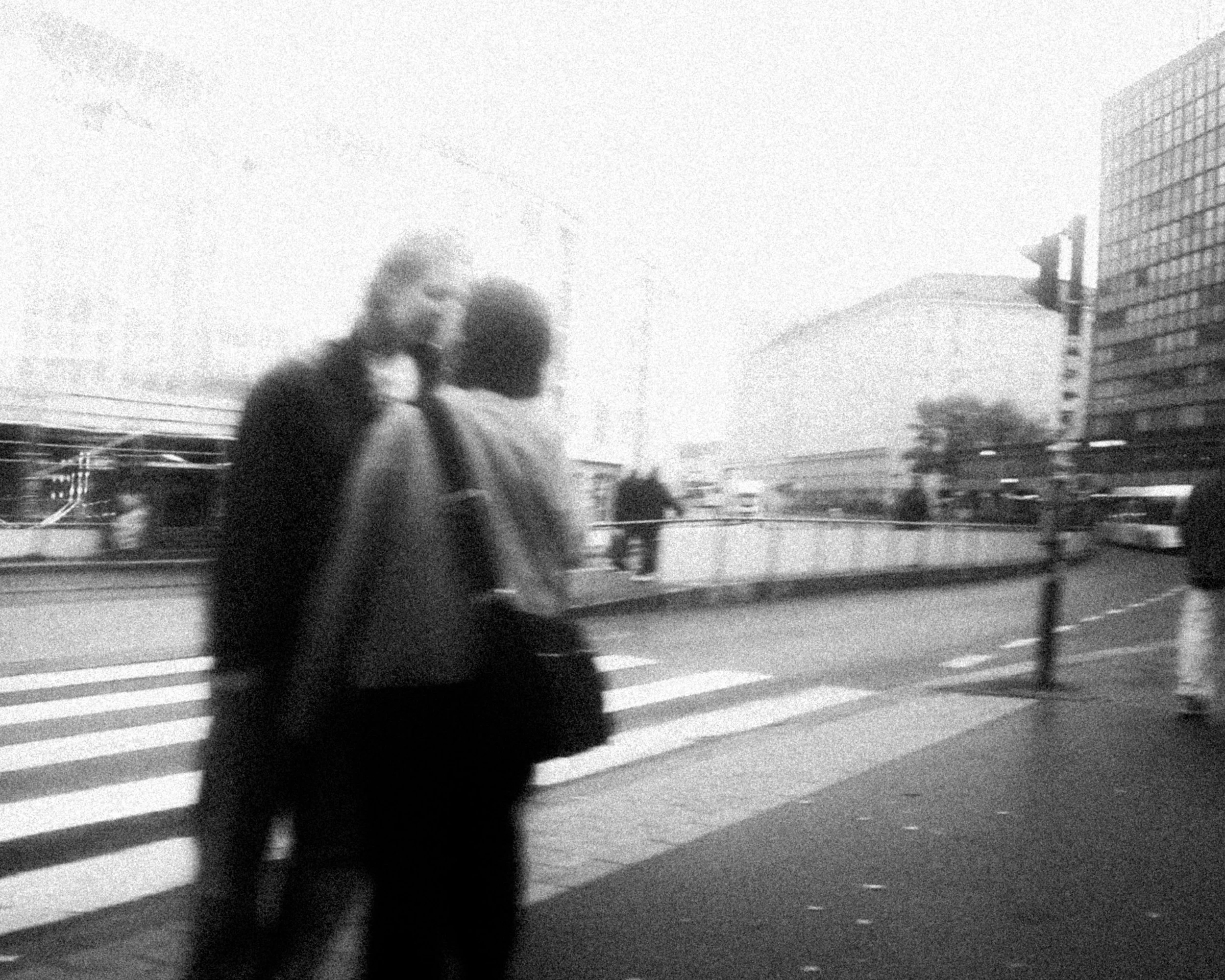Black and white photo of two people walking across a city street at a crosswalk, with urban buildings and a bus in the background.