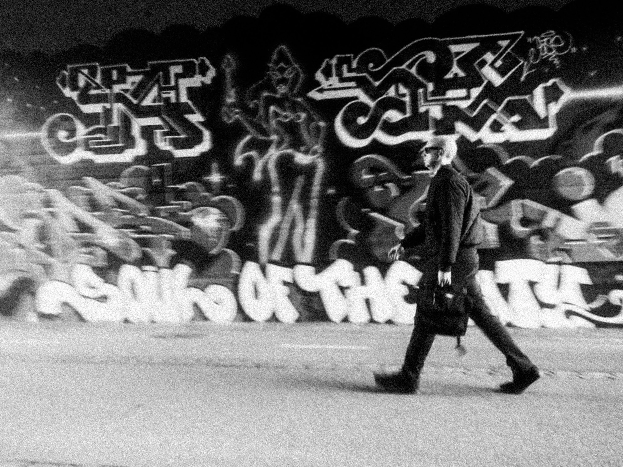 A man dressed in dark clothing with sunglasses and a cap walking past a colorful graffiti mural at night.