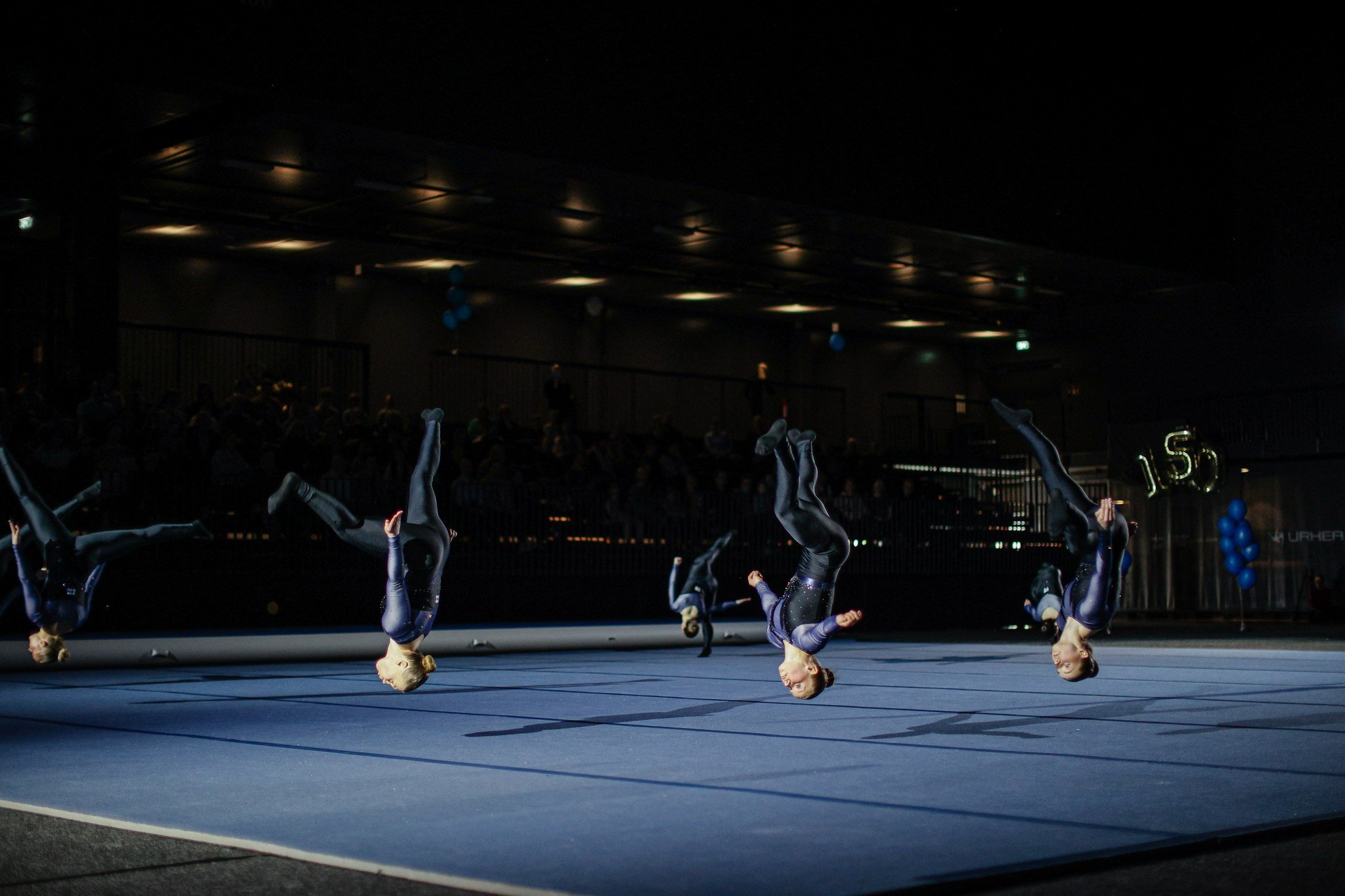 Gymnasts performing flips on a blue mat in a dark indoor arena with audience in the background, some holding balloons.