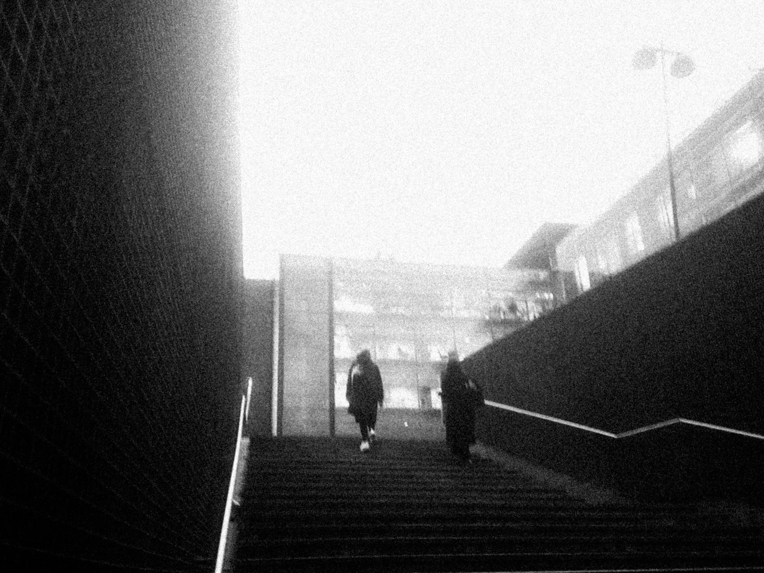 Two people walk up a set of outdoor stairs near tall buildings on a foggy day.