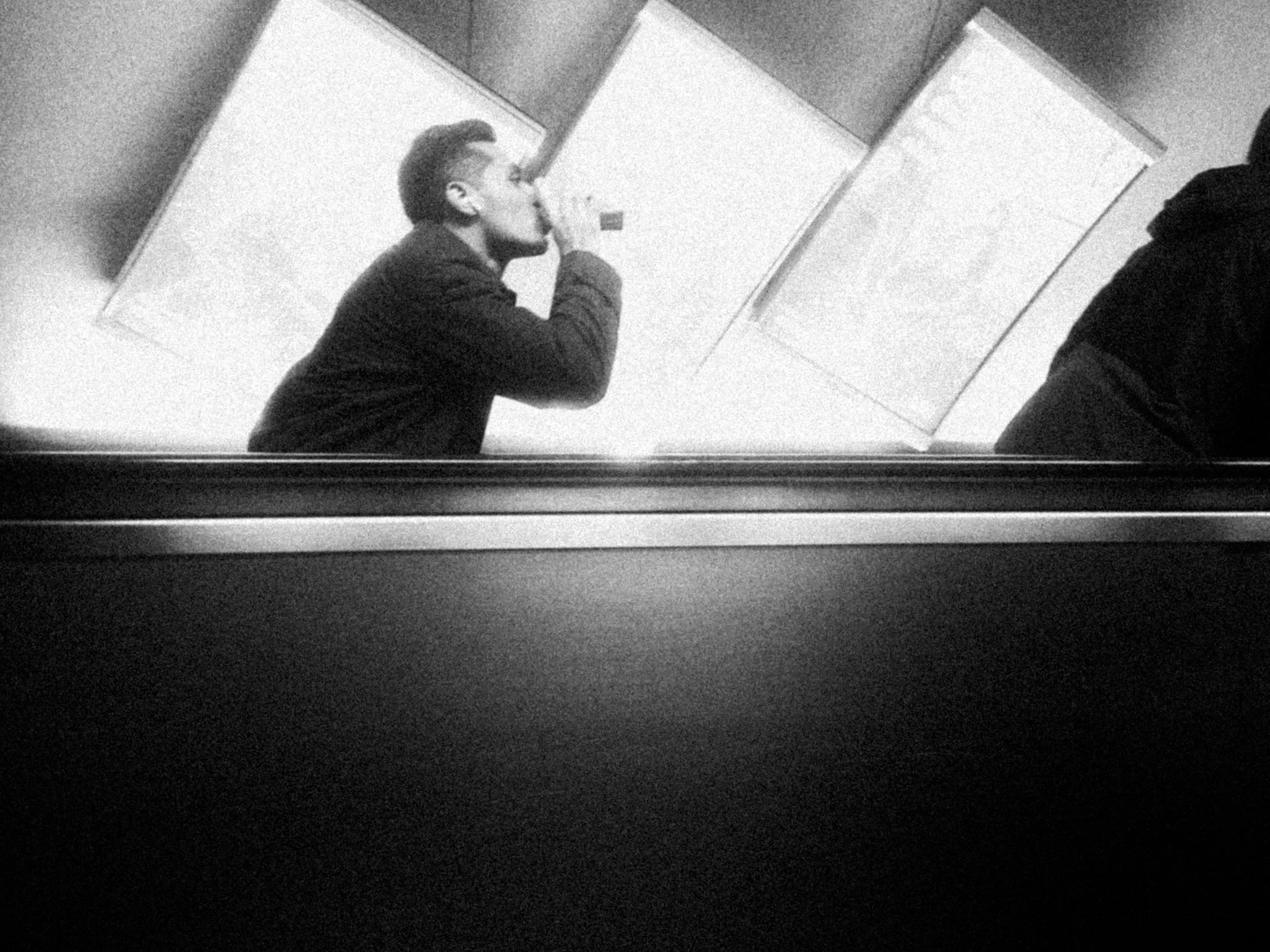 A young man in a dark jacket is sitting at a desk or counter, holding a cup or mug close to his face, possibly drinking from it. The setting appears to be indoors, and the photo is in black and white.