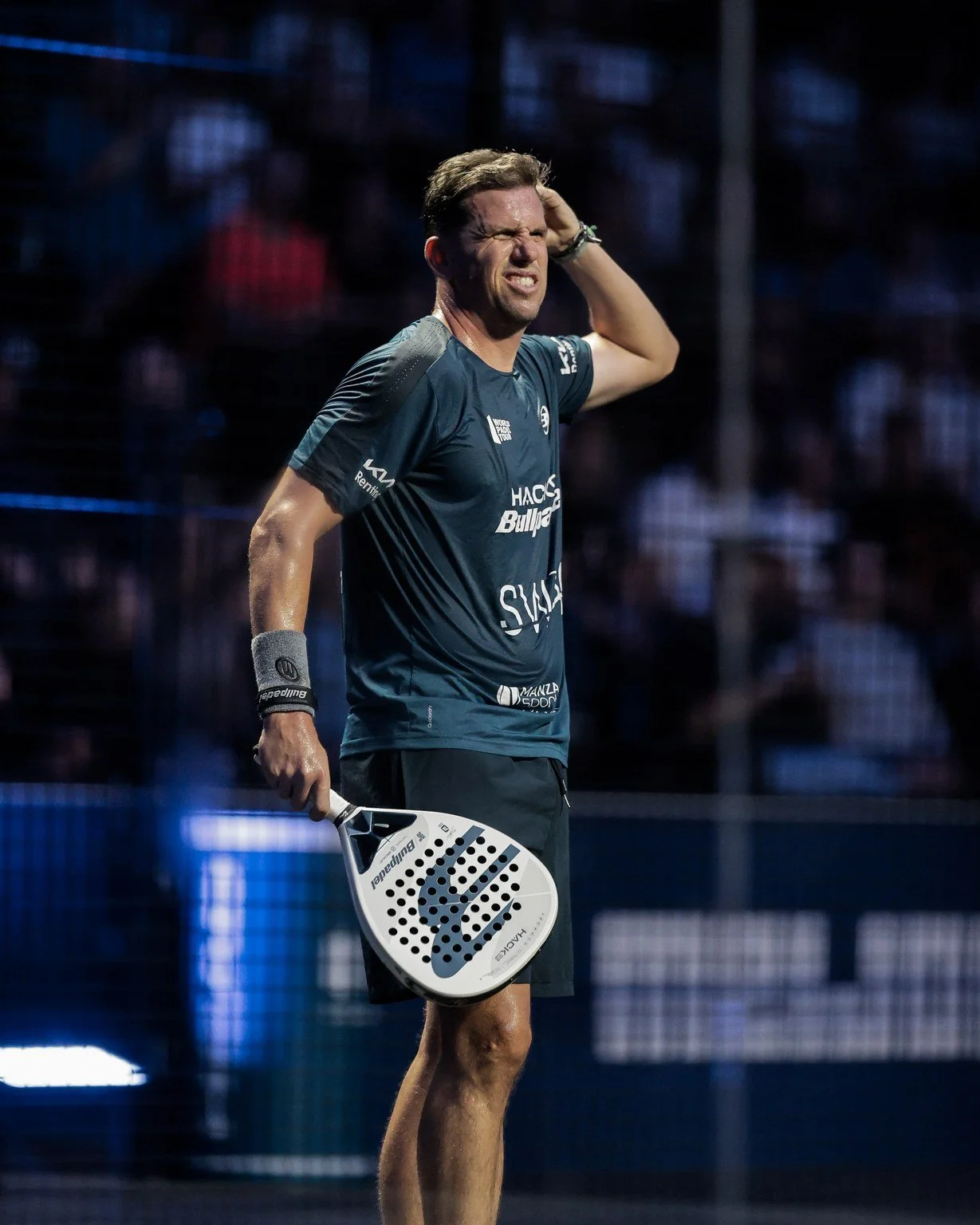 A male padel player standing on a court holding a padel racket, with a frustrated or disappointed expression, scratching his head.