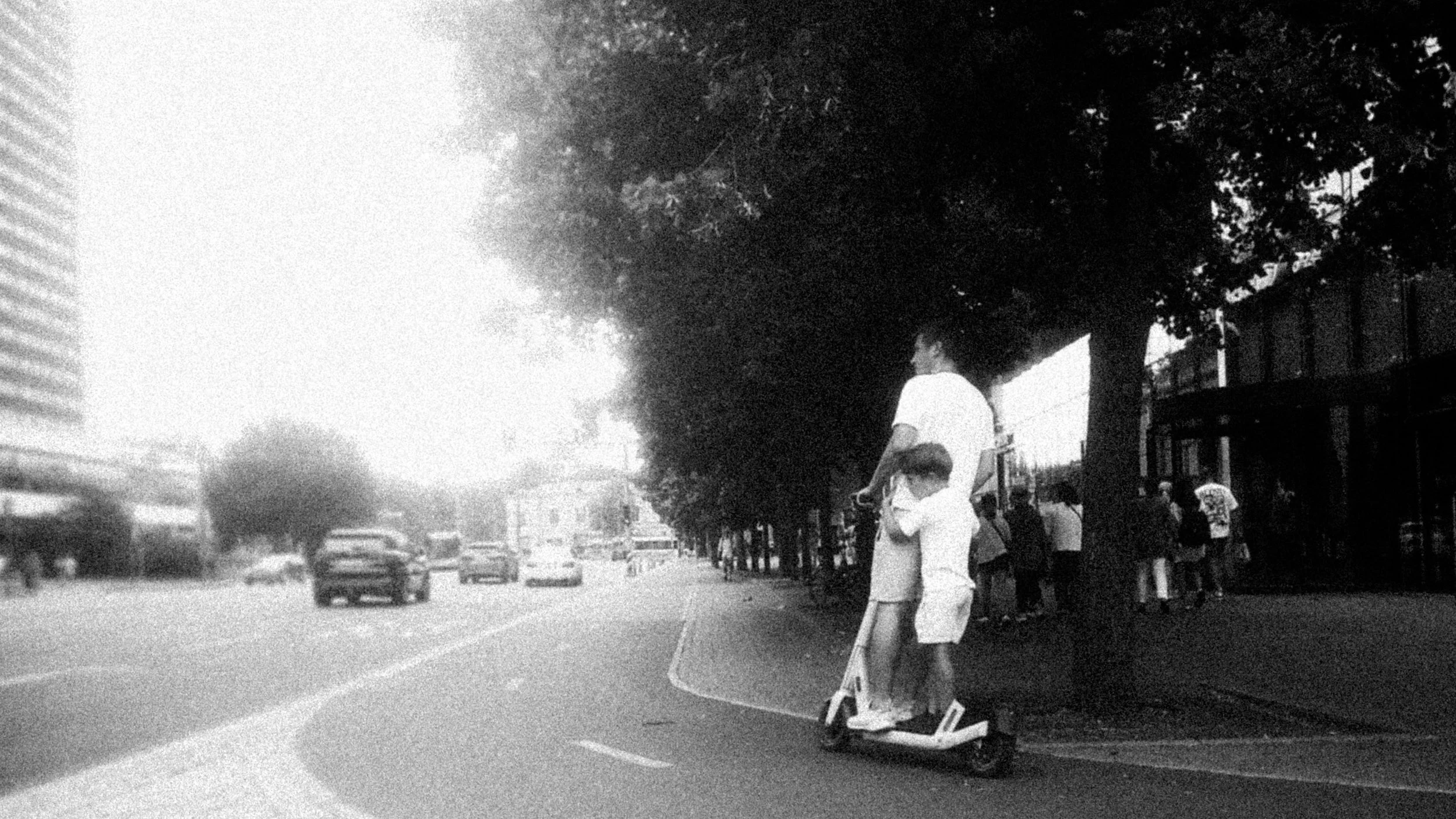 A person riding an electric scooter on a city sidewalk near a row of trees and a building, with other pedestrians in the background.
