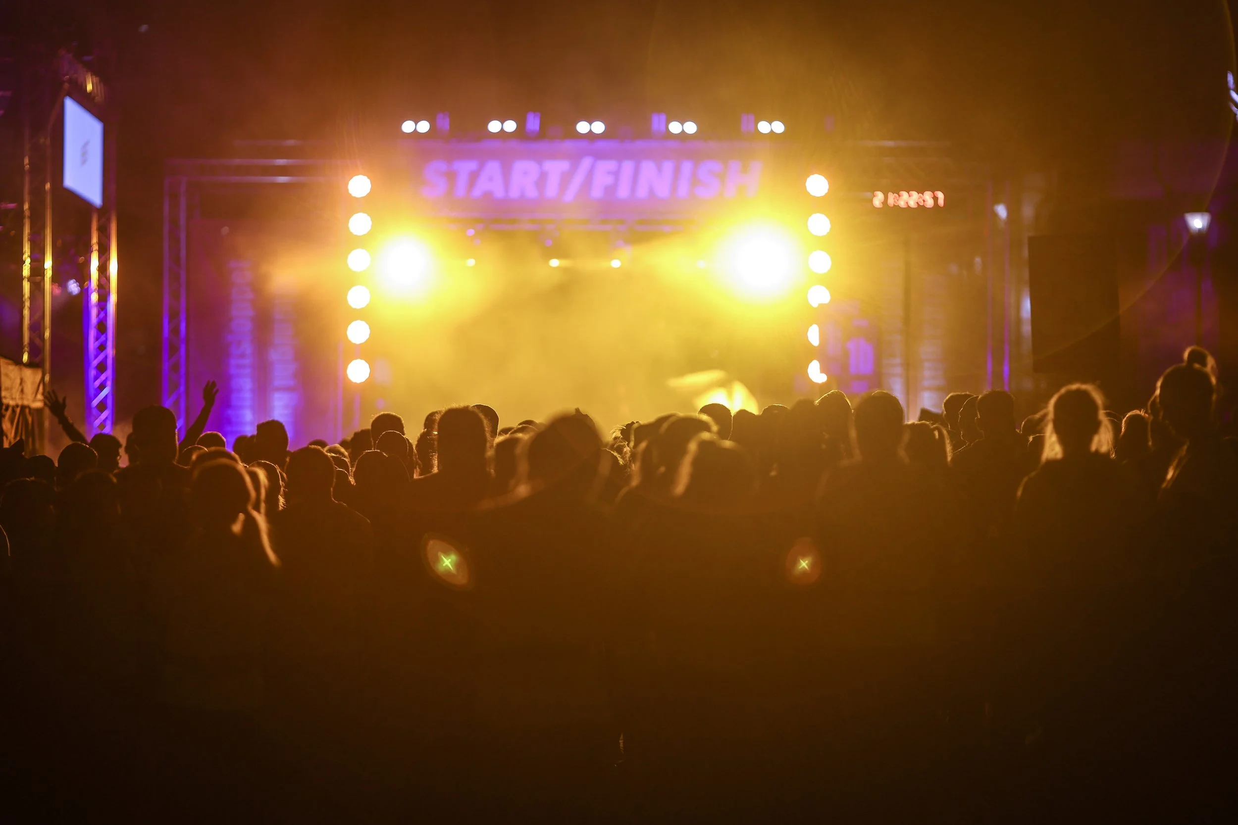 Crowd gathered at a concert or event with bright yellow stage lights and a 'Start/Finish' sign, possibly at a race or festival.