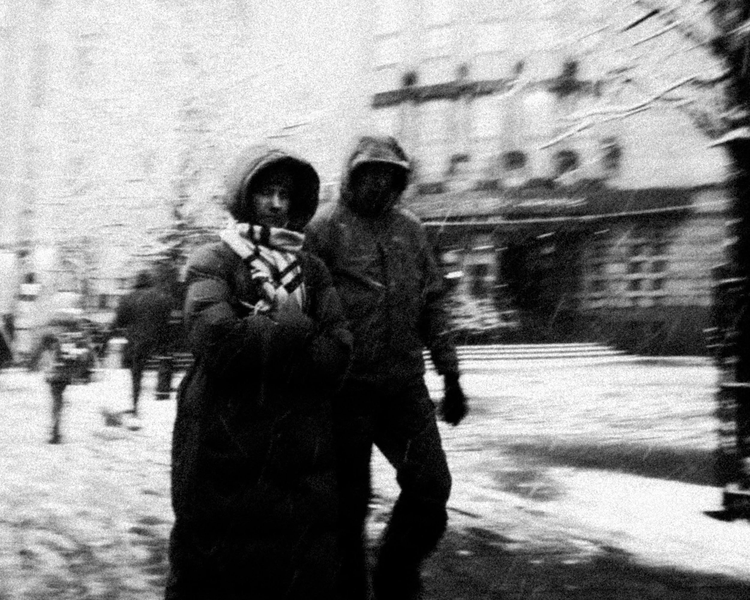 Black and white photograph of two children walking in a city street during winter, wearing warm jackets and hats, with blurred buildings and people in the background.